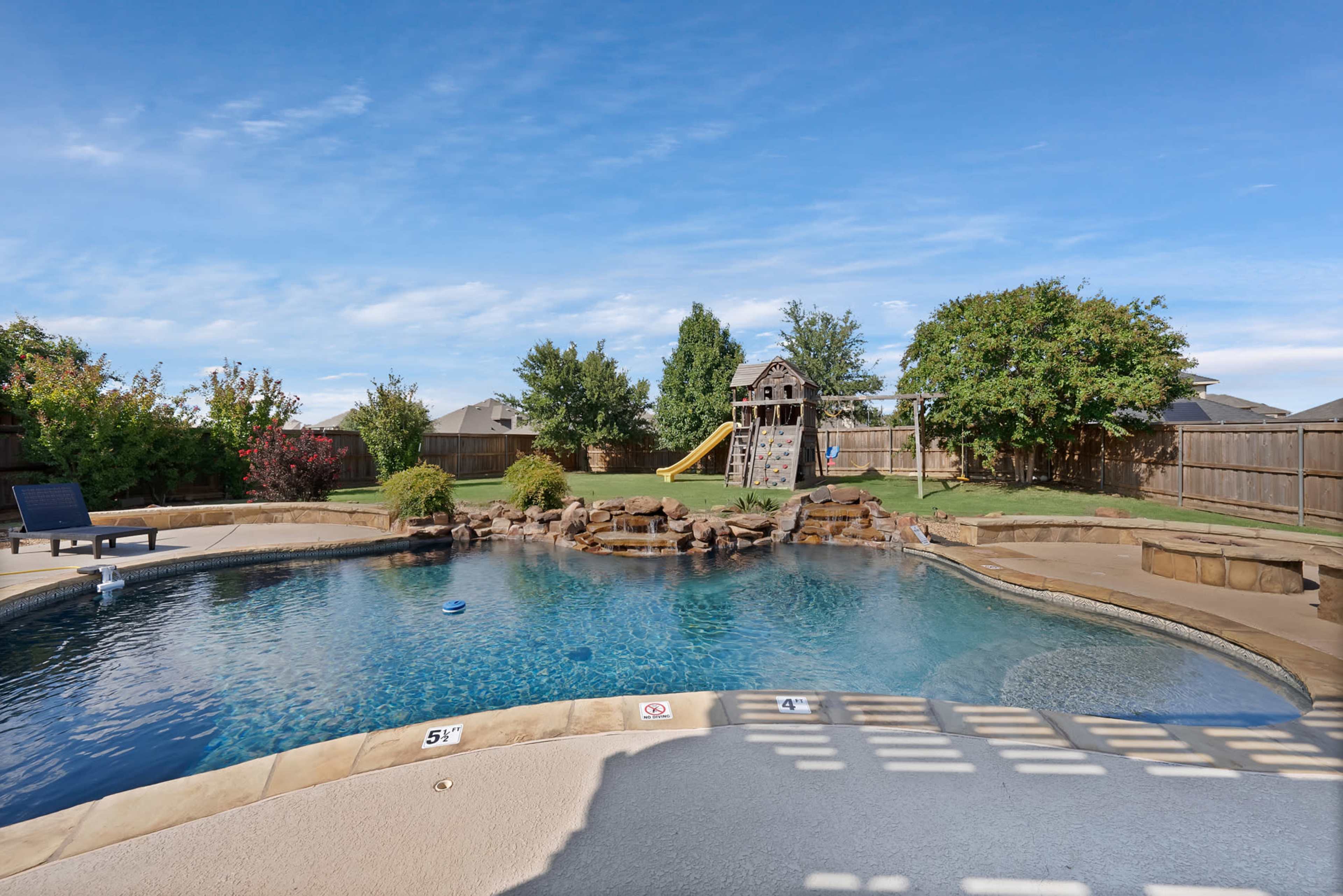 The image depicts a swimming pool with a stone waterfall, surrounded by a grassy yard and a playground set in the background.