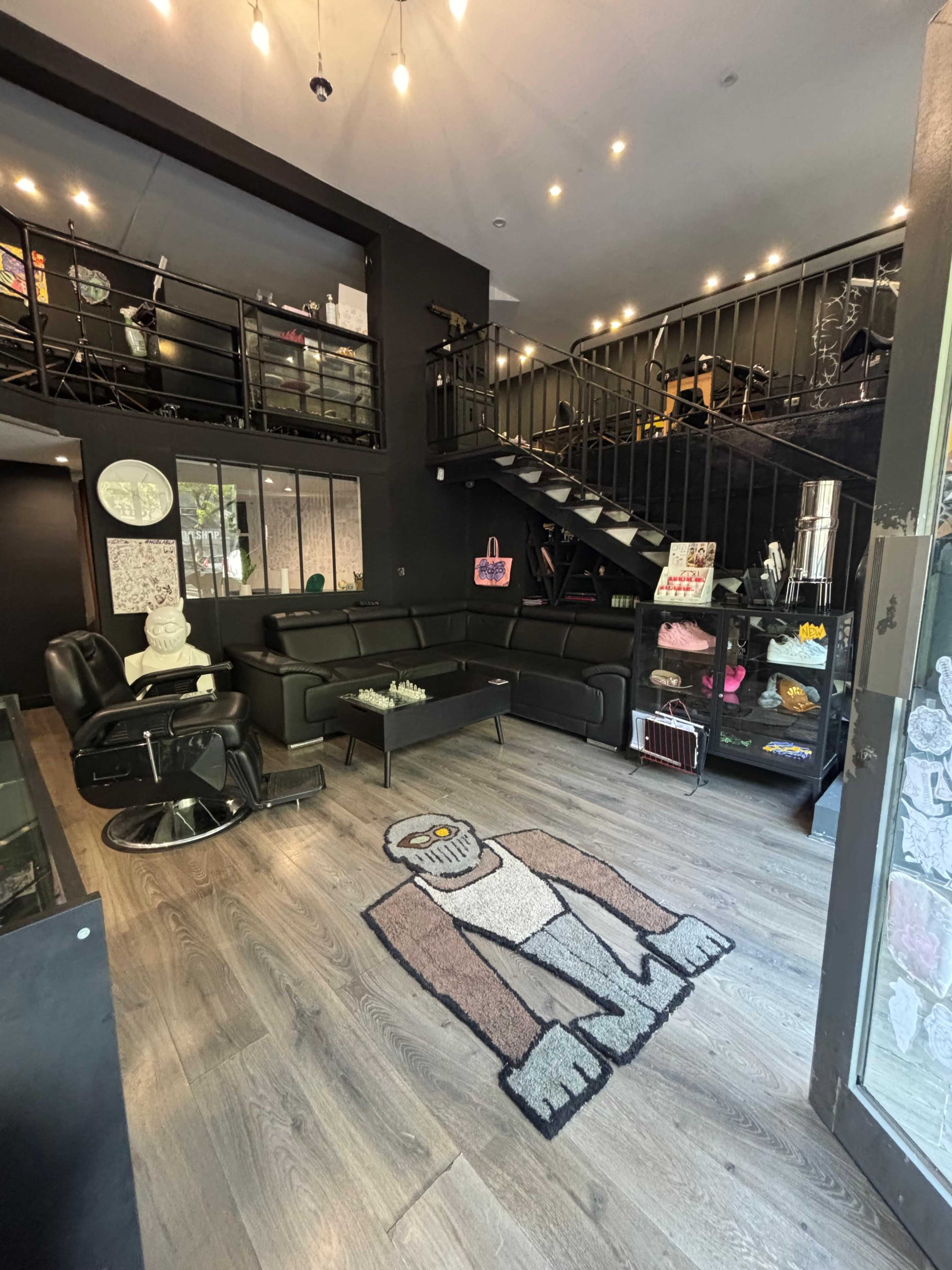 The image shows a modern, two-level interior of a black-themed café or lounge, featuring a staircase, seating area with black couches, and decorative elements.
