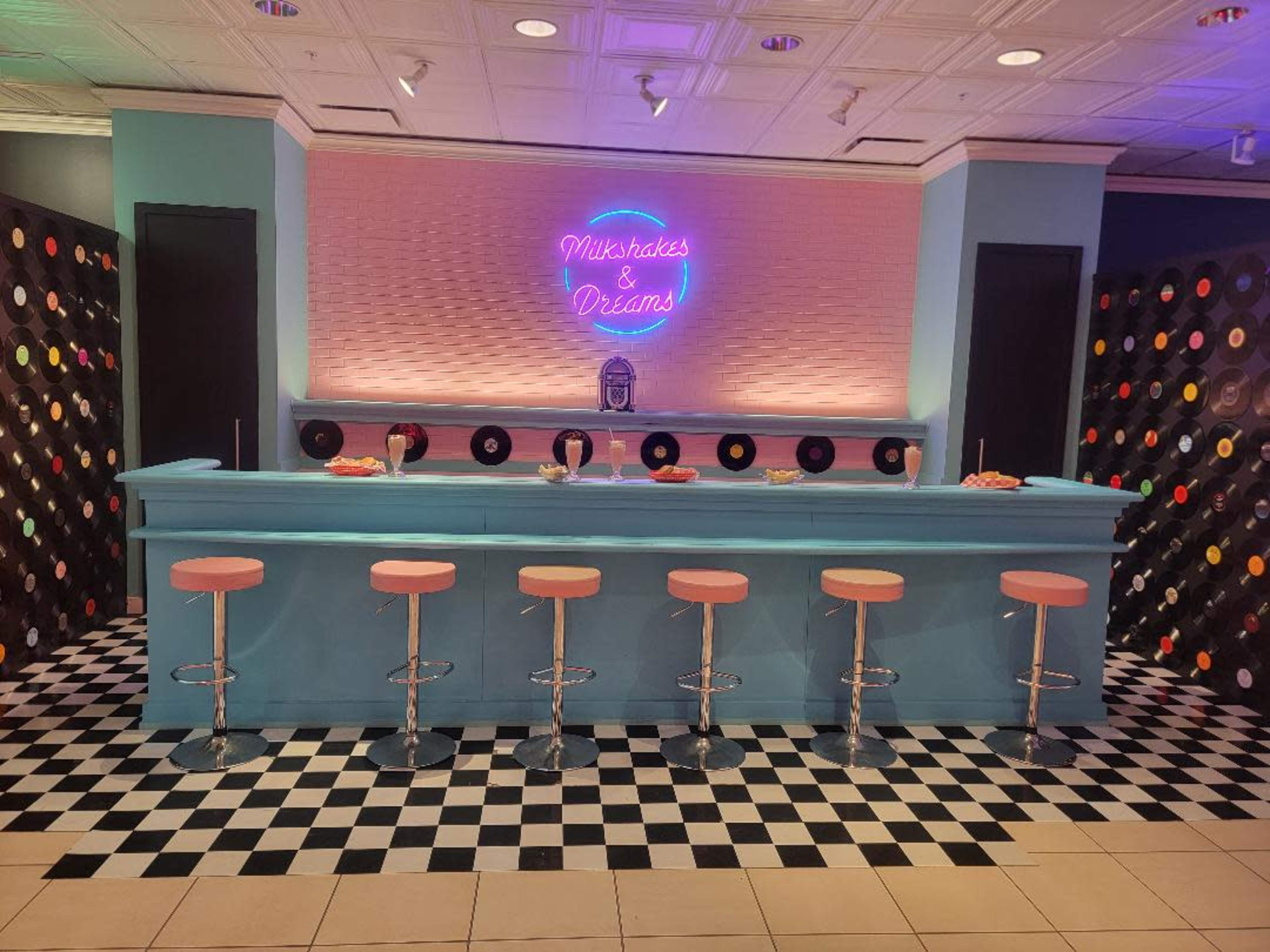 A retro-style diner with a light blue counter, pink stools, and a neon sign that reads "Milkshakes & Dreams," set against a patterned black and white tiled floor.