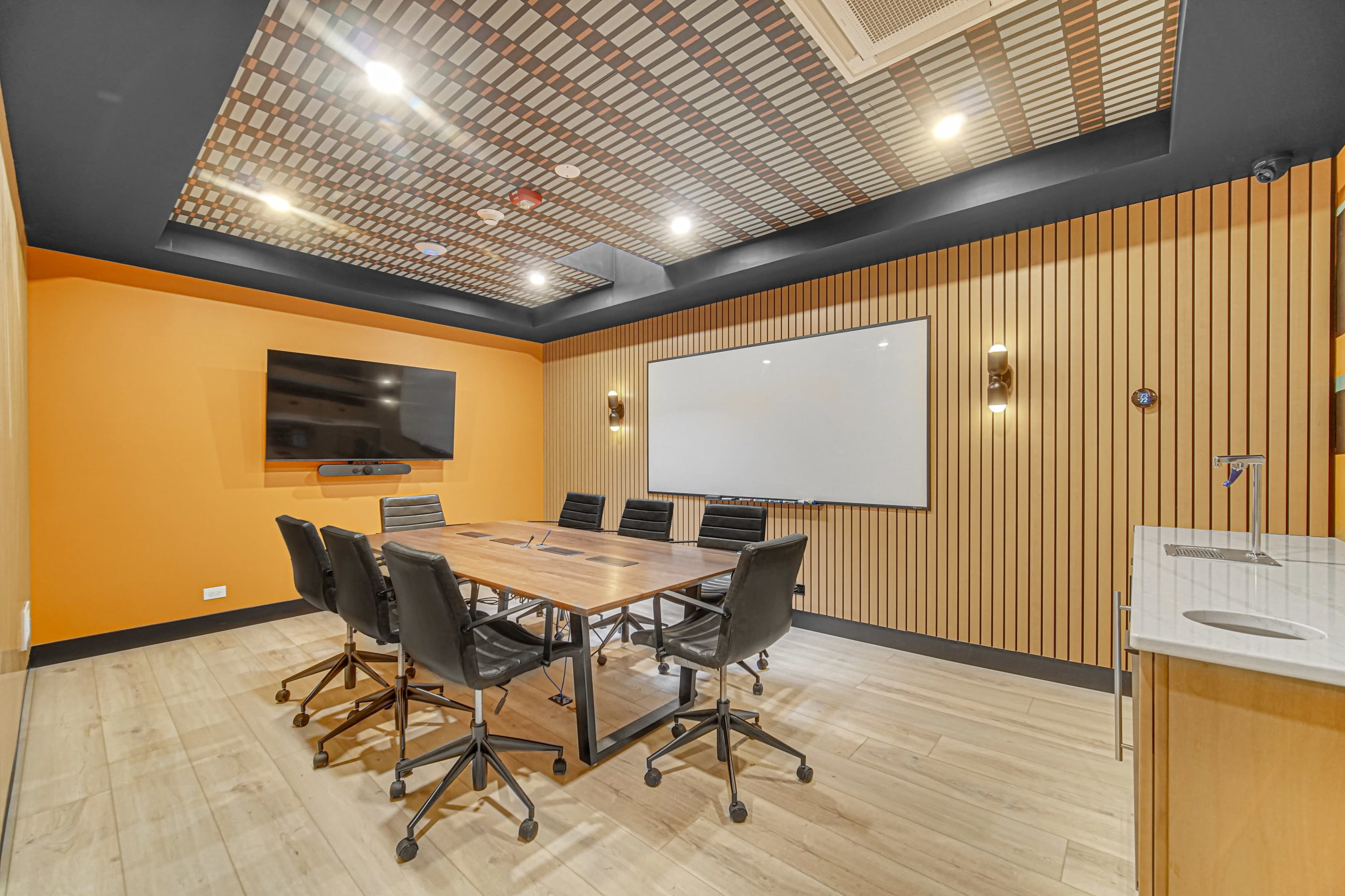 The image shows a modern conference room with a large wooden table, black chairs, a wall-mounted TV, and an orange accent wall, featuring a whiteboard and stylish ceiling design.