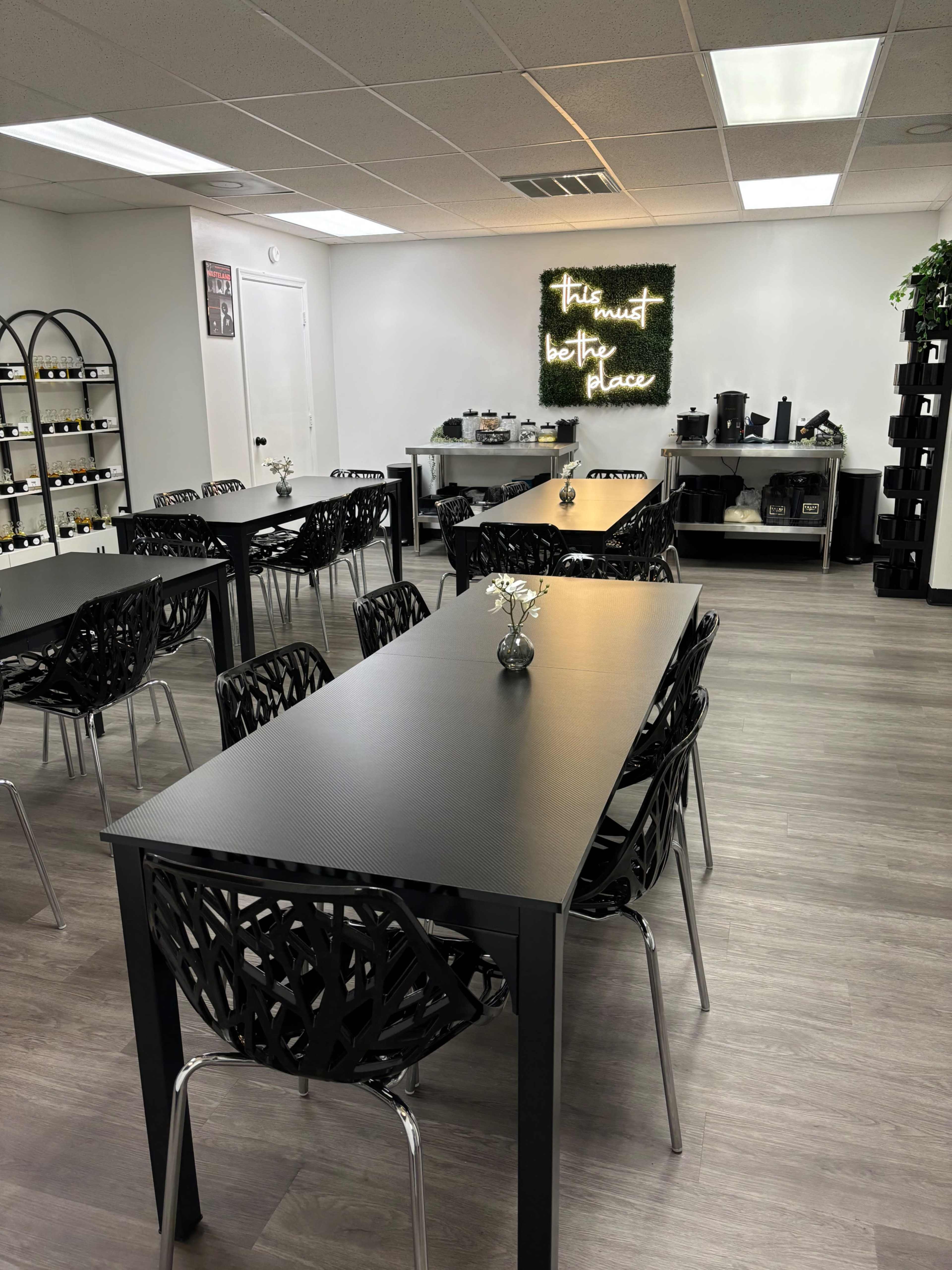 A modern, well-lit room with several black tables and chairs arranged neatly, a wall with decorative lettering, and a counter with coffee and tea supplies.