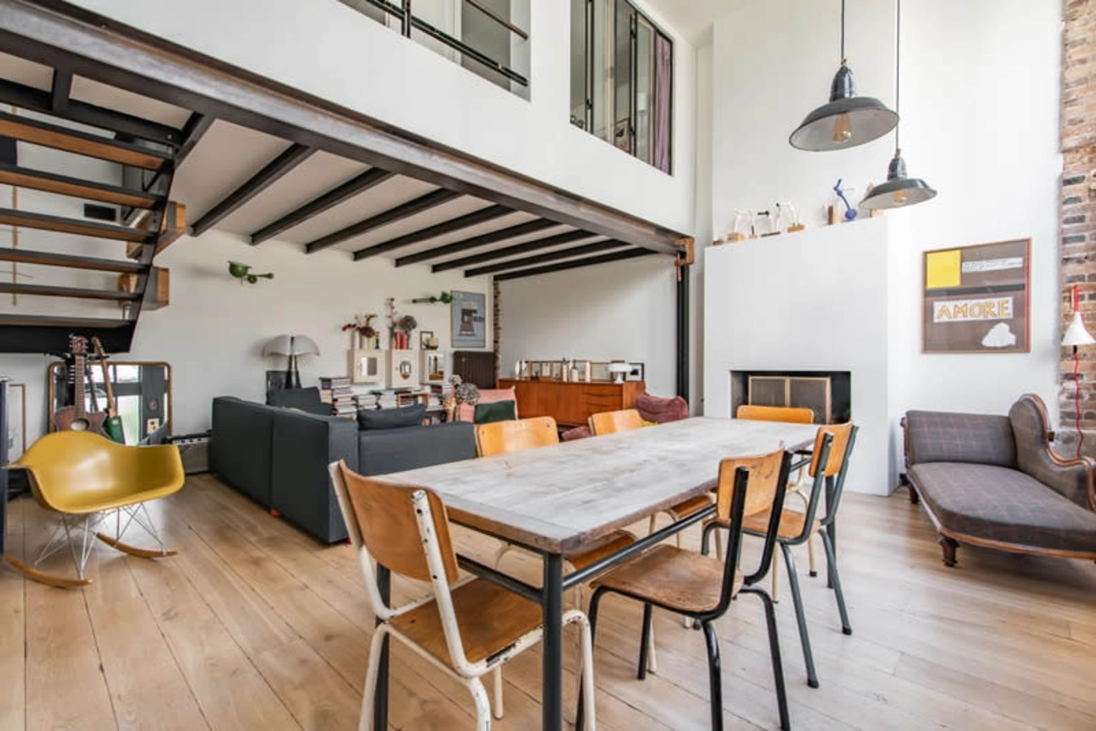 A spacious living area featuring a large wooden dining table surrounded by metal and wooden chairs, with a seating area visible in the background and a staircase leading to an upper level.