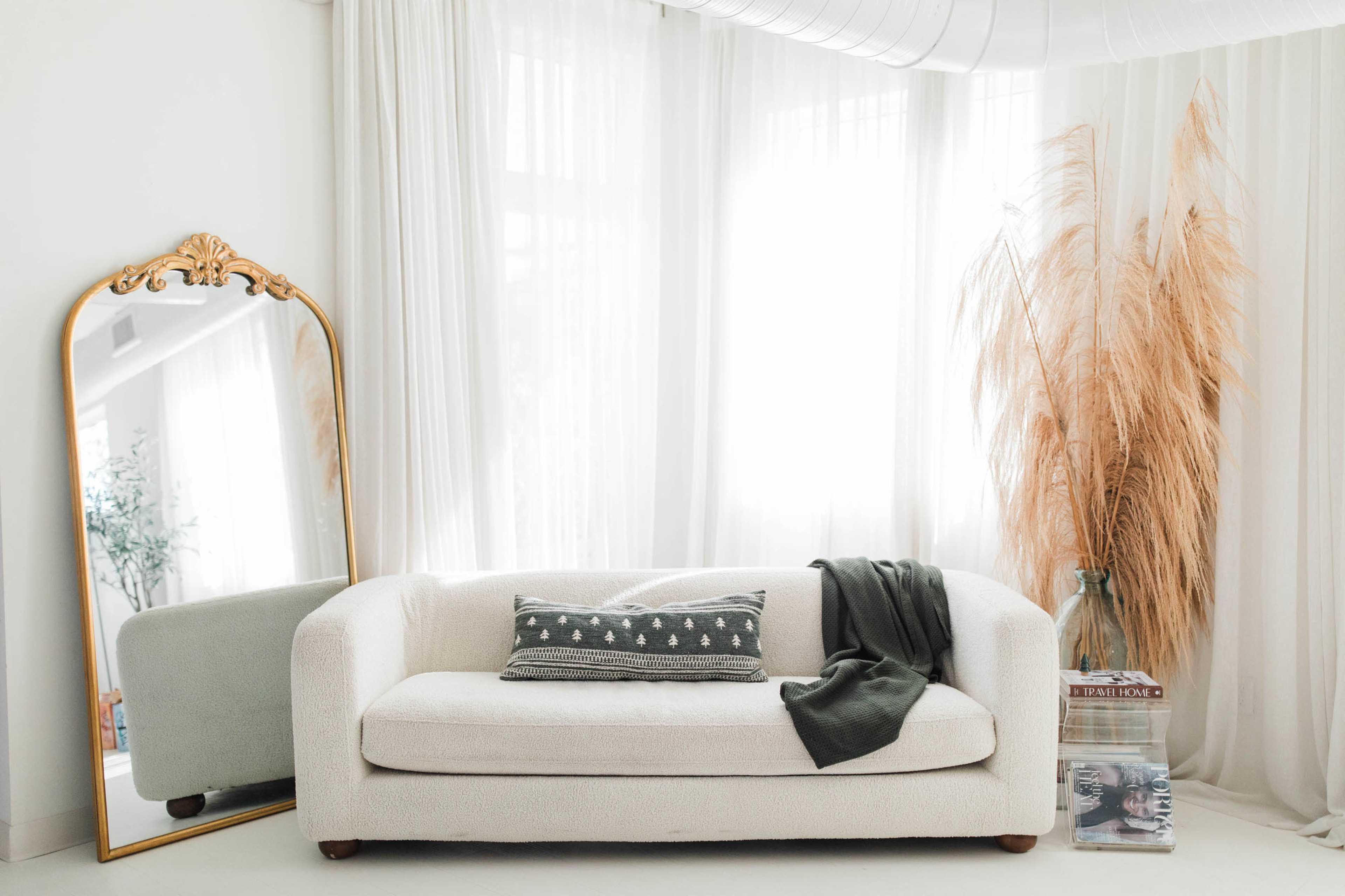A light-colored sofa with a decorative pillow sits beside a tall mirror and a vase near a large window with flowing curtains.