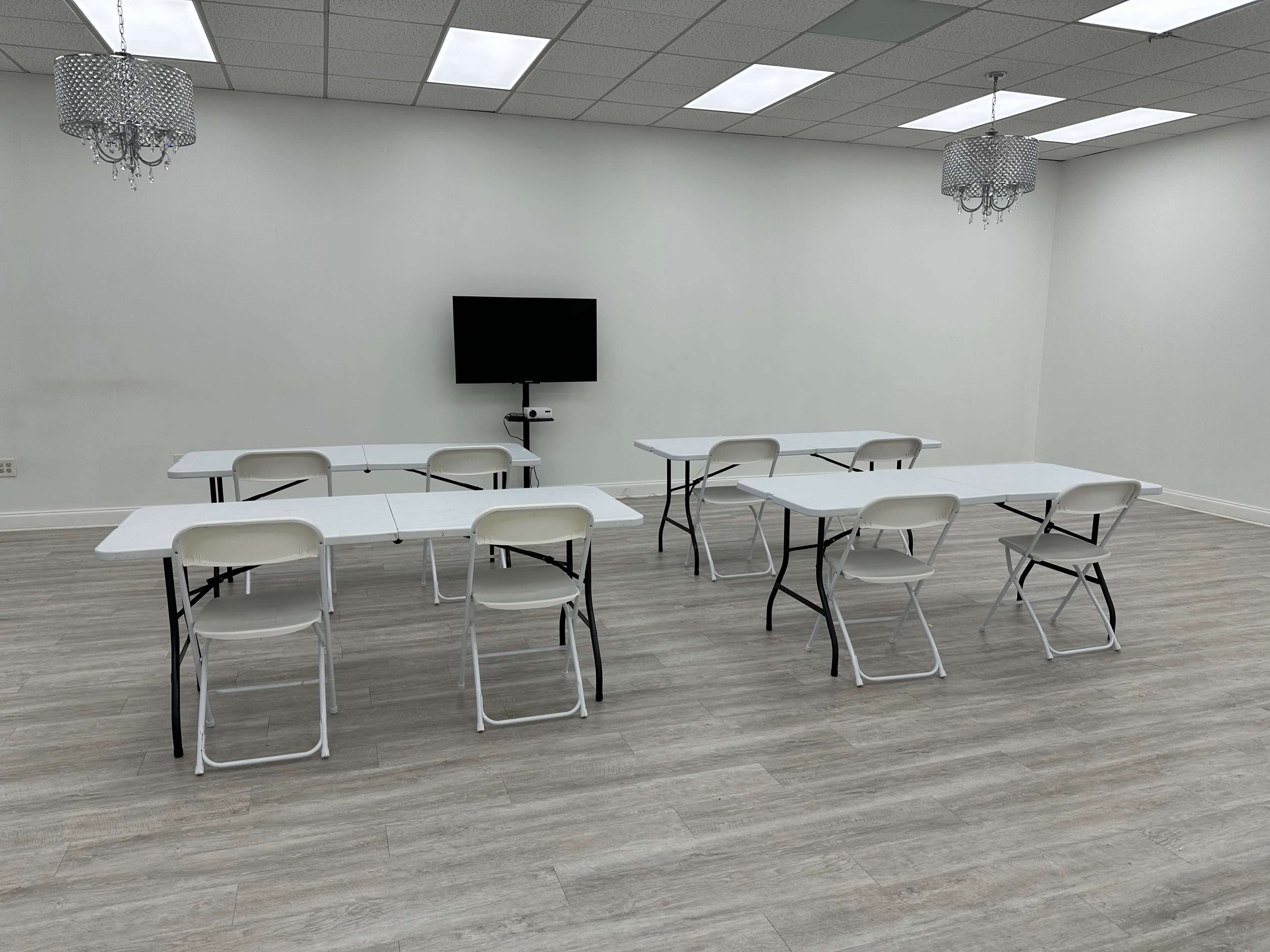A simple, empty room features several folding tables and chairs arranged for a meeting, with a television mounted on the wall.