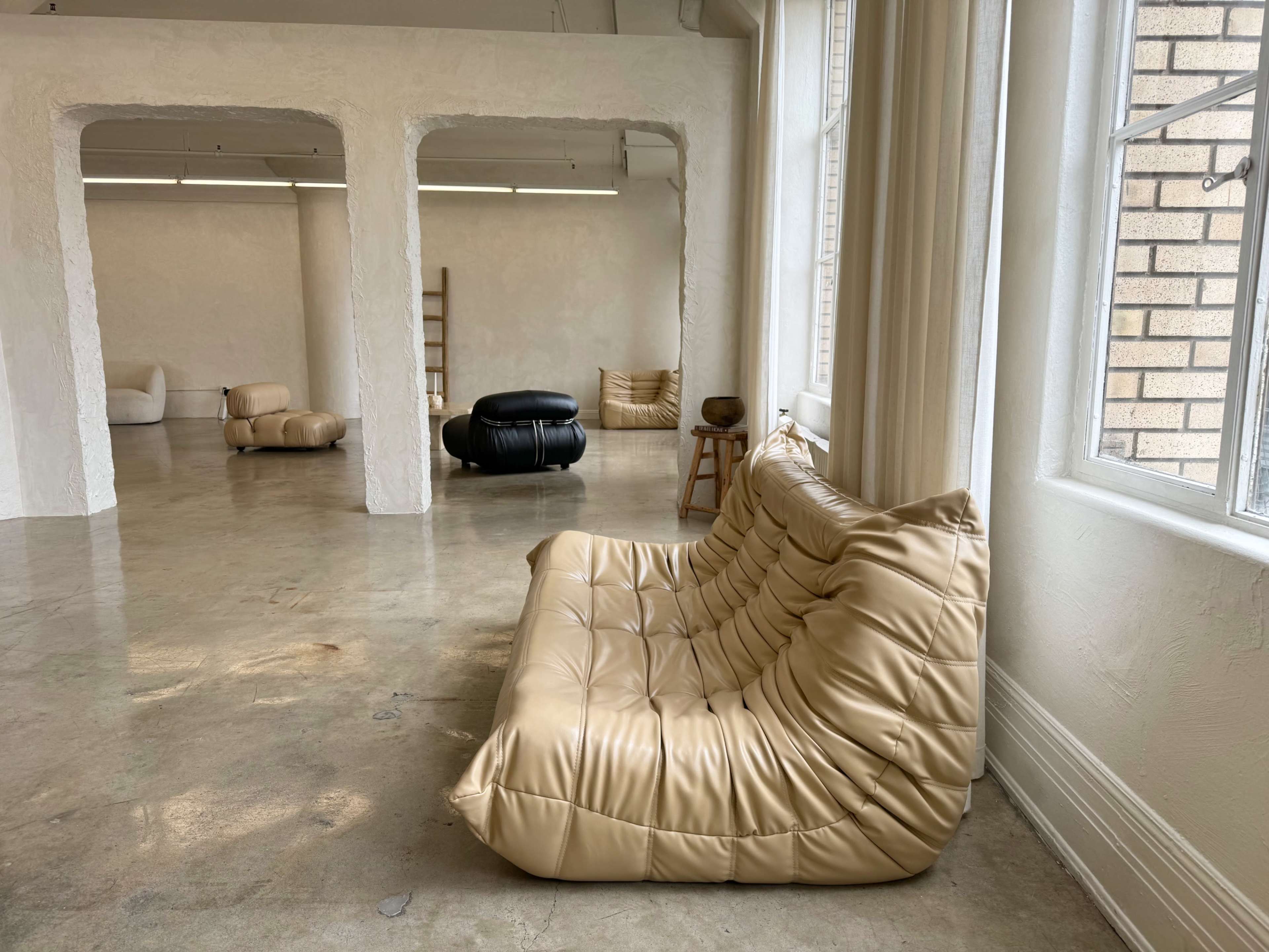 A spacious, minimalist room featuring a beige, padded sofa, several minimalist furniture pieces, and large windows allowing natural light to enter.