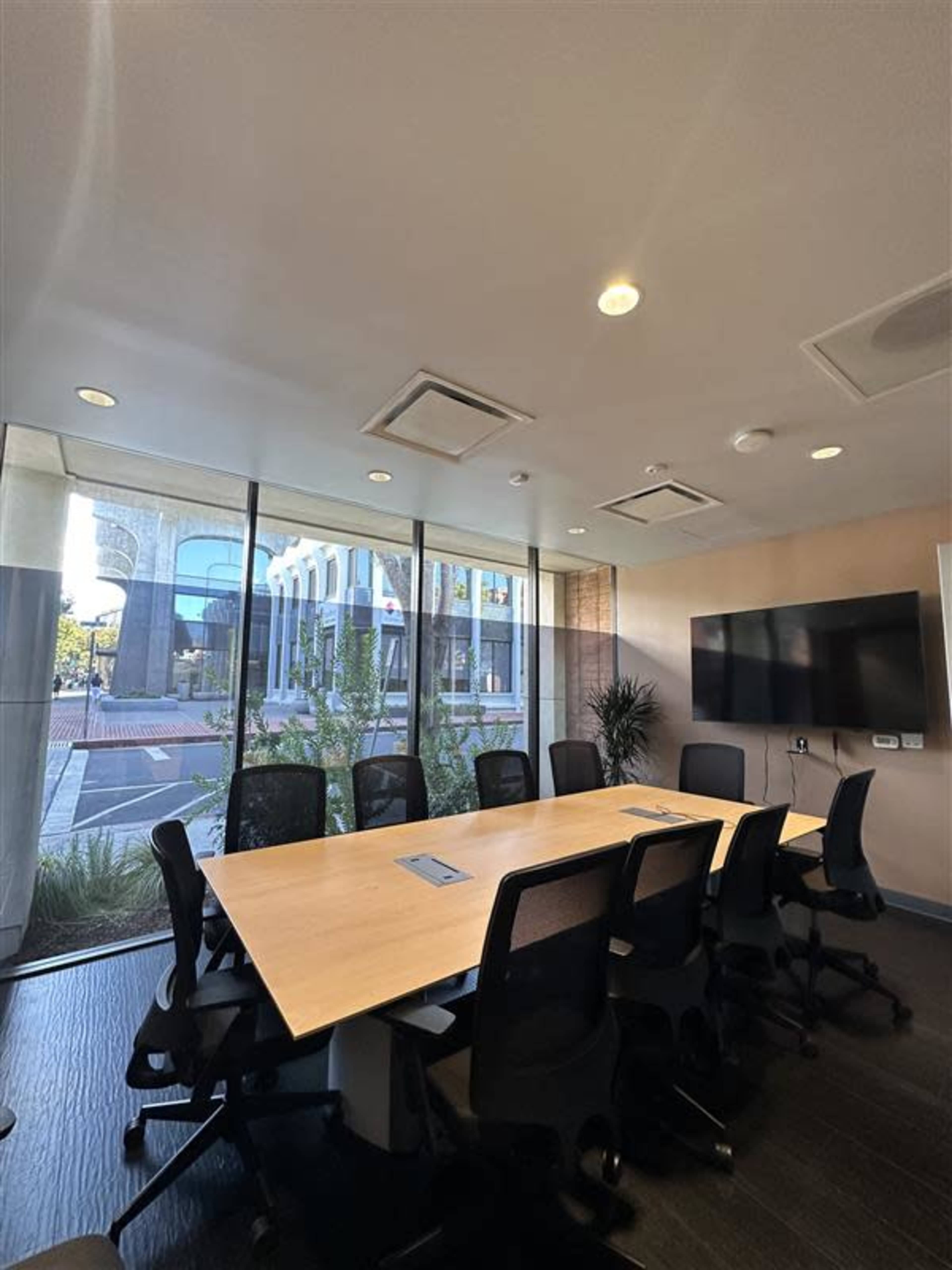 A modern conference room with a large wooden table, ergonomic chairs, and floor-to-ceiling windows displaying a view of an outdoor plaza.