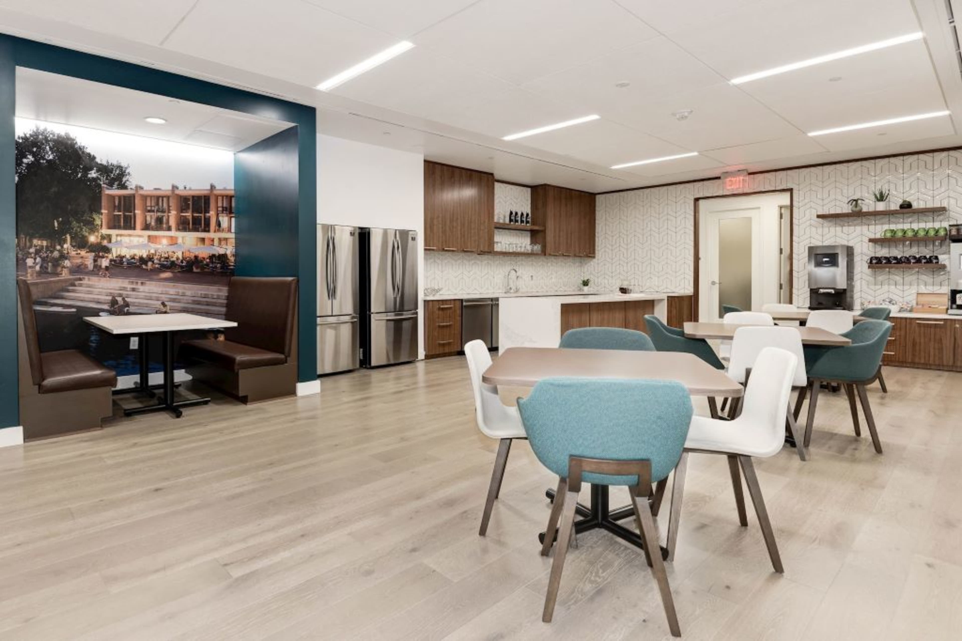 The image shows a modern kitchenette area with seating, featuring a large mural on the wall, stainless steel appliances, and a patterned backsplash.