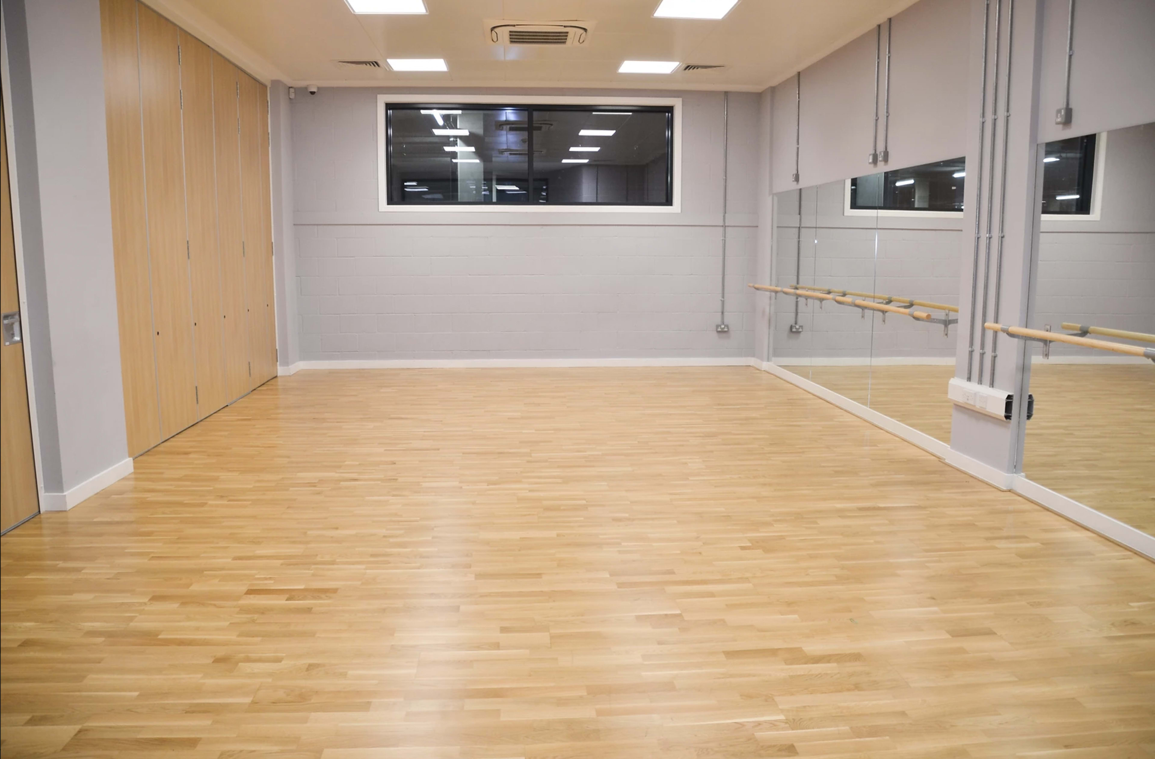 An empty dance studio with wooden flooring, large mirrors, and a window.