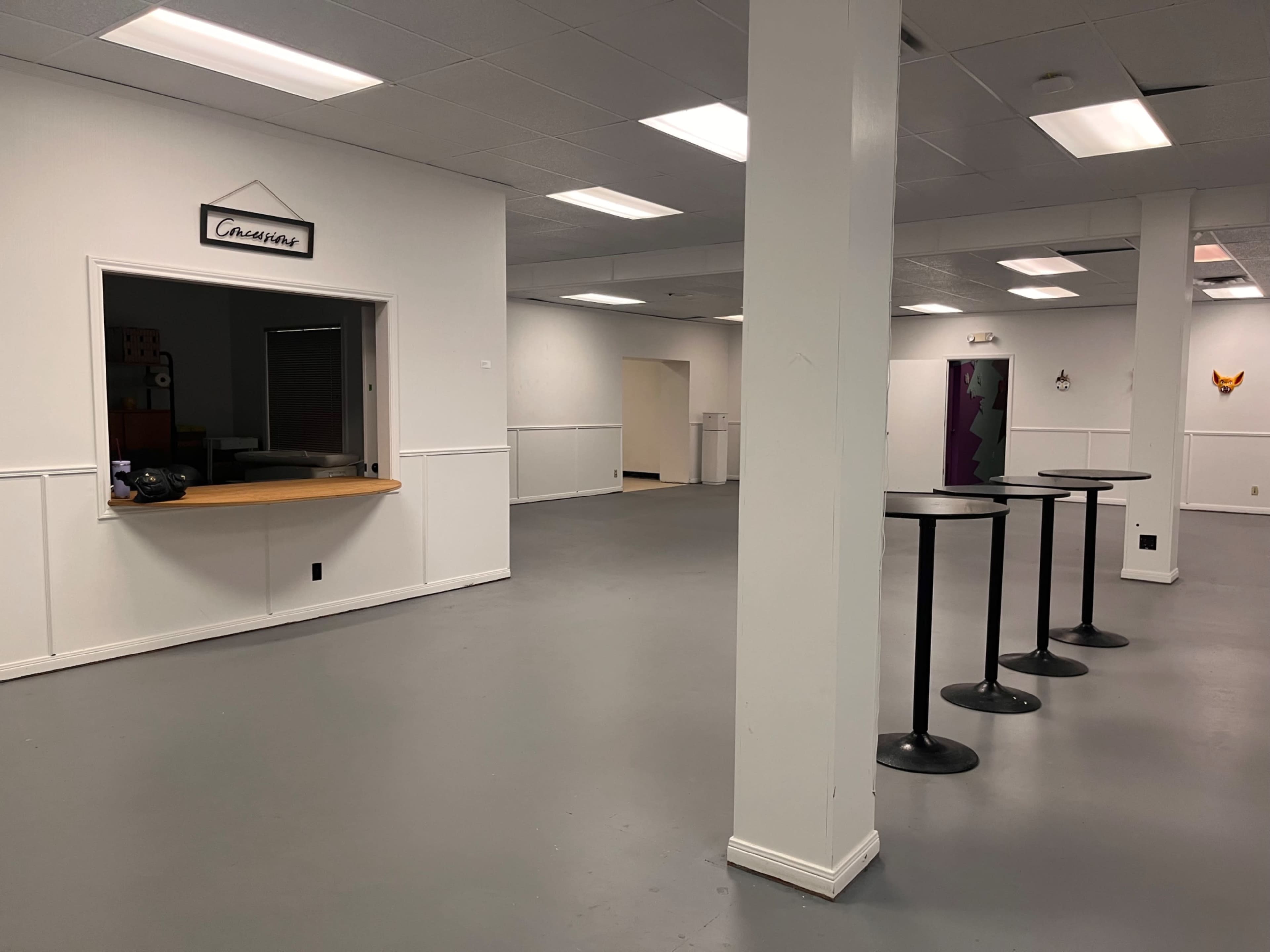 The image shows a spacious, empty room with white walls, a service window marked "Canteen," and several tall, round tables arranged throughout.