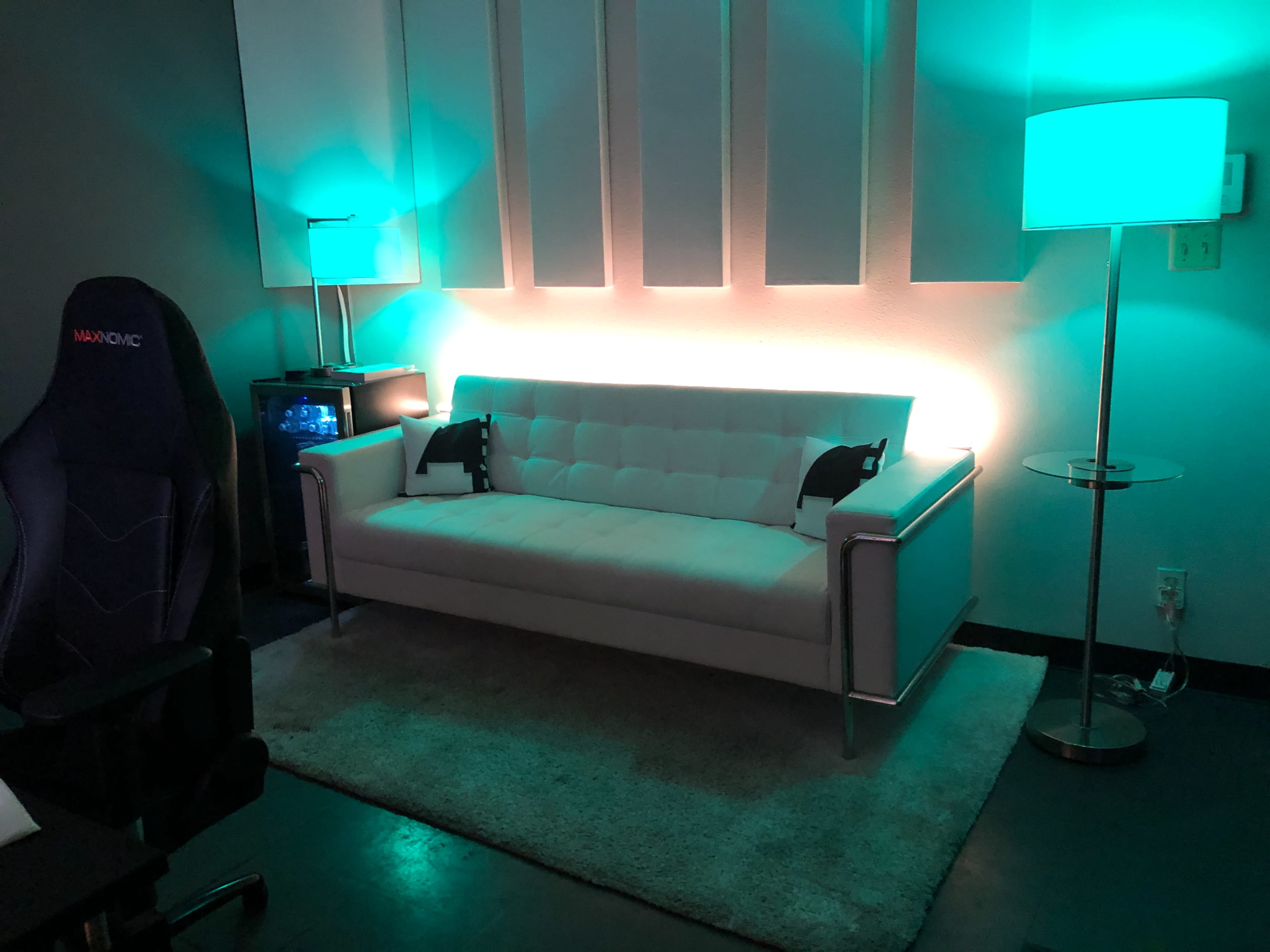 A modern living room setup featuring a white couch, a side table with a lamp, and a gaming chair, all illuminated by teal lighting.