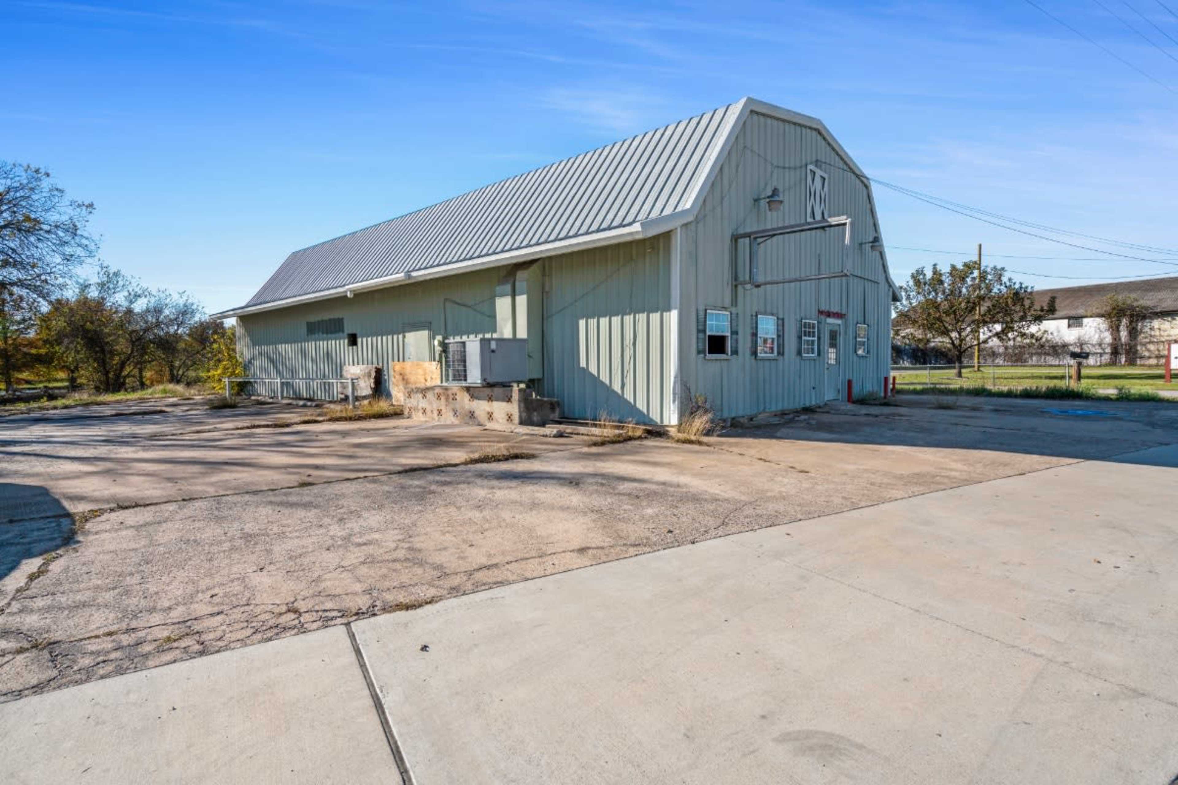 A single-story metal building with a peaked roof stands beside a paved area and is surrounded by sparse landscaping.