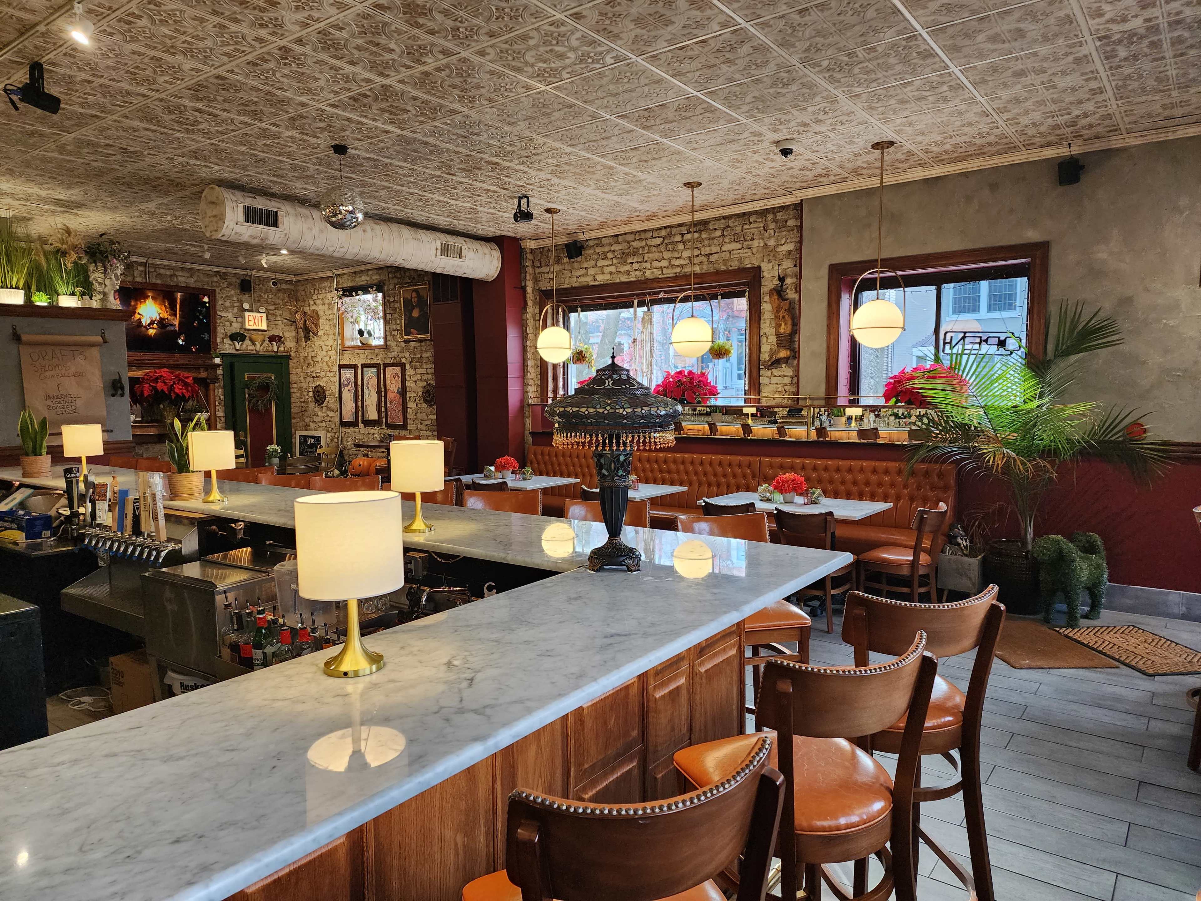 The image shows the interior of a restaurant featuring a marble bar with brass lamps, wooden seating, and decor including plants and red floral arrangements.