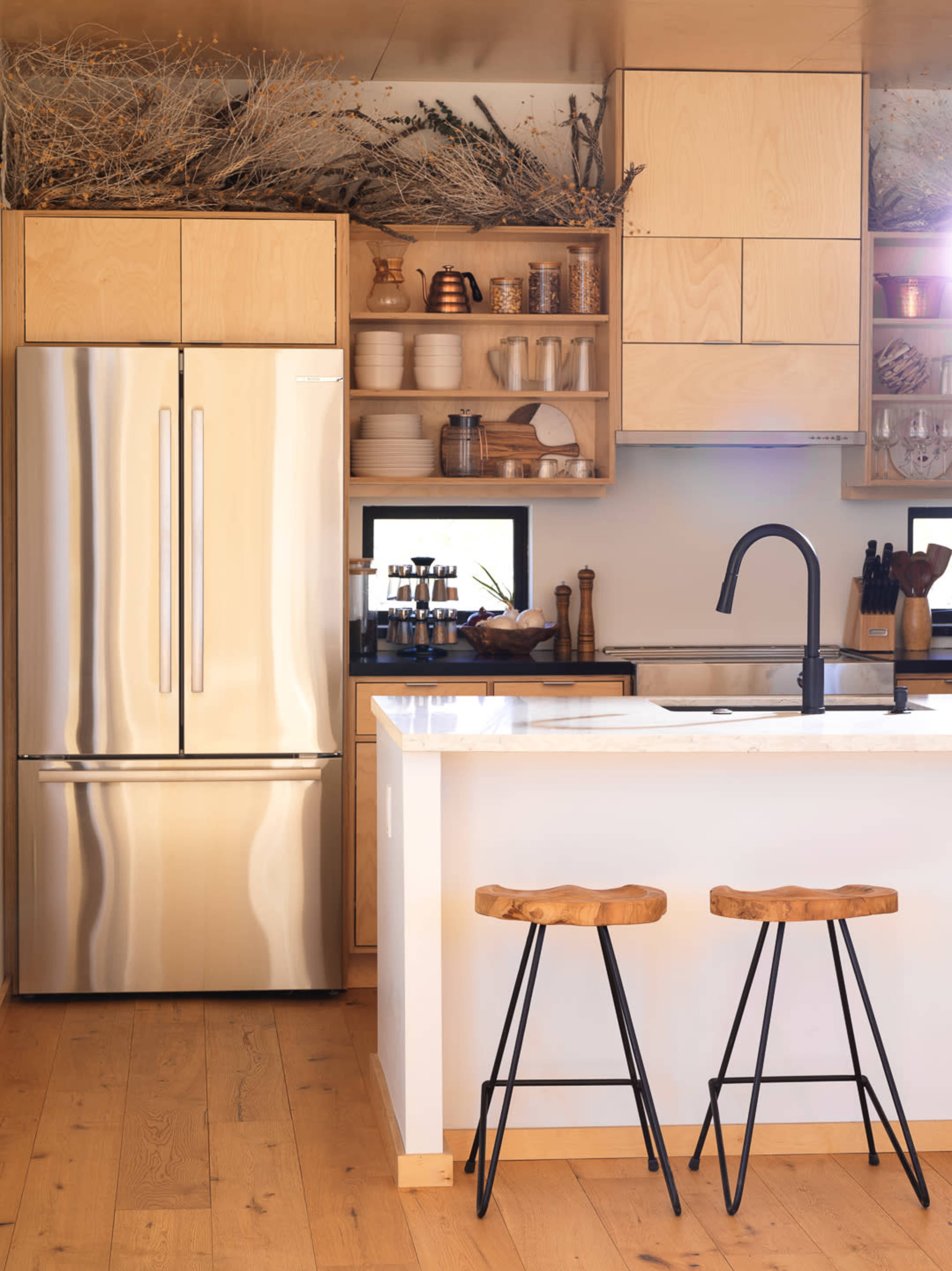 A modern kitchen features a stainless steel refrigerator, light wooden cabinets, open shelving with dishware, and two metal-legged stools at a white island.