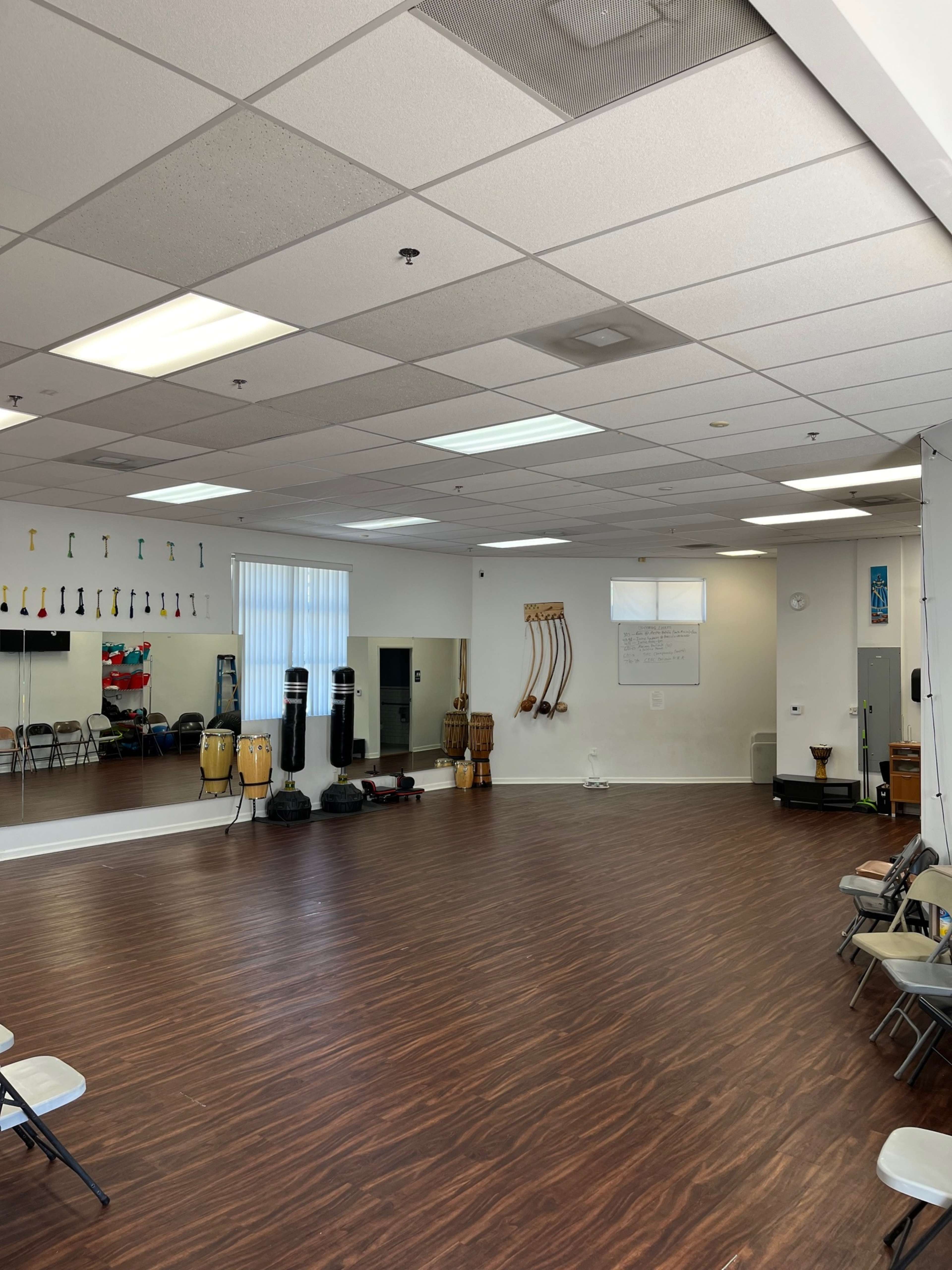 The image shows a spacious dance or exercise studio with wooden flooring, mirrors on one wall, and various percussion instruments and chairs arranged around the room.