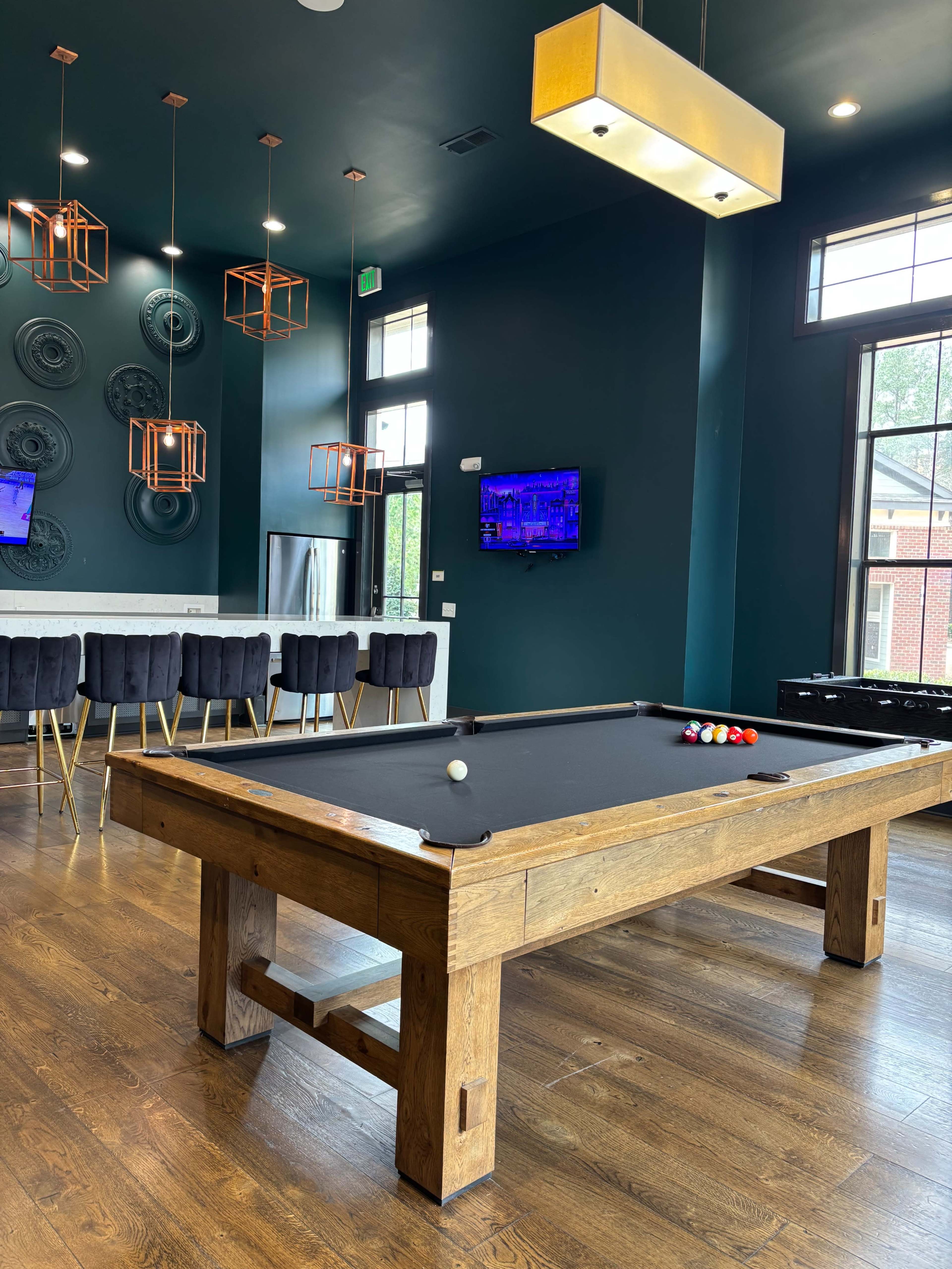 The image shows a spacious lounge area featuring a wooden pool table, modern lighting fixtures, and a bar with high-backed chairs.