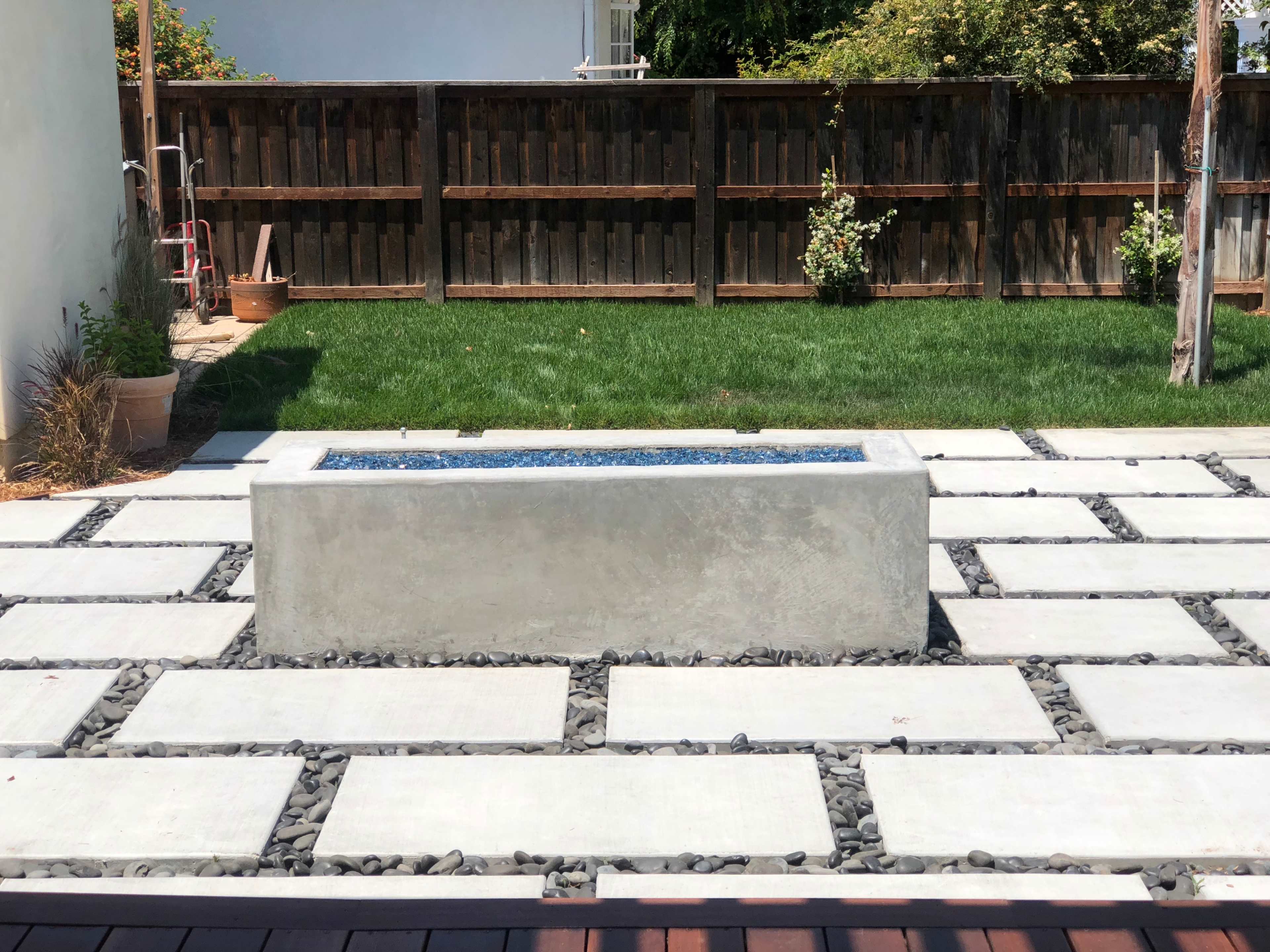 The image shows a rectangular concrete planter filled with blue stones, situated on a patio made of concrete slabs and pebbles, with a grassy area and wooden fence in the background.