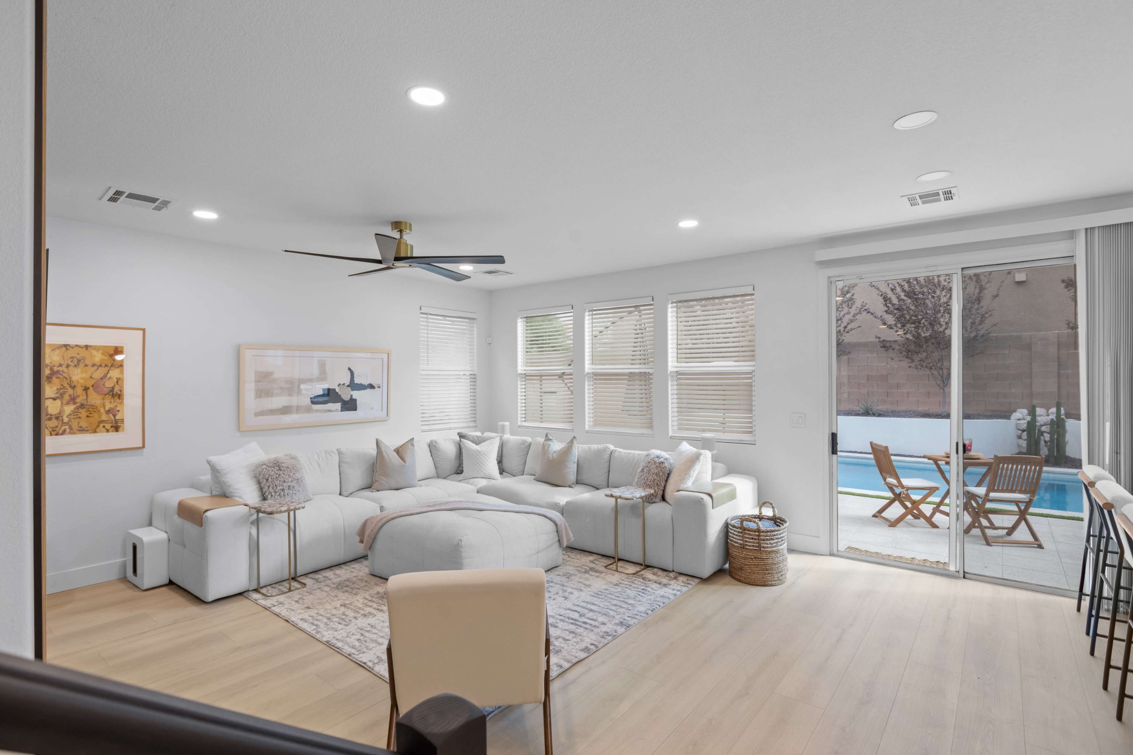 The image shows a modern living room with a large sectional couch, a coffee table, and sliding glass doors that lead to an outdoor area featuring a pool and patio seating.
