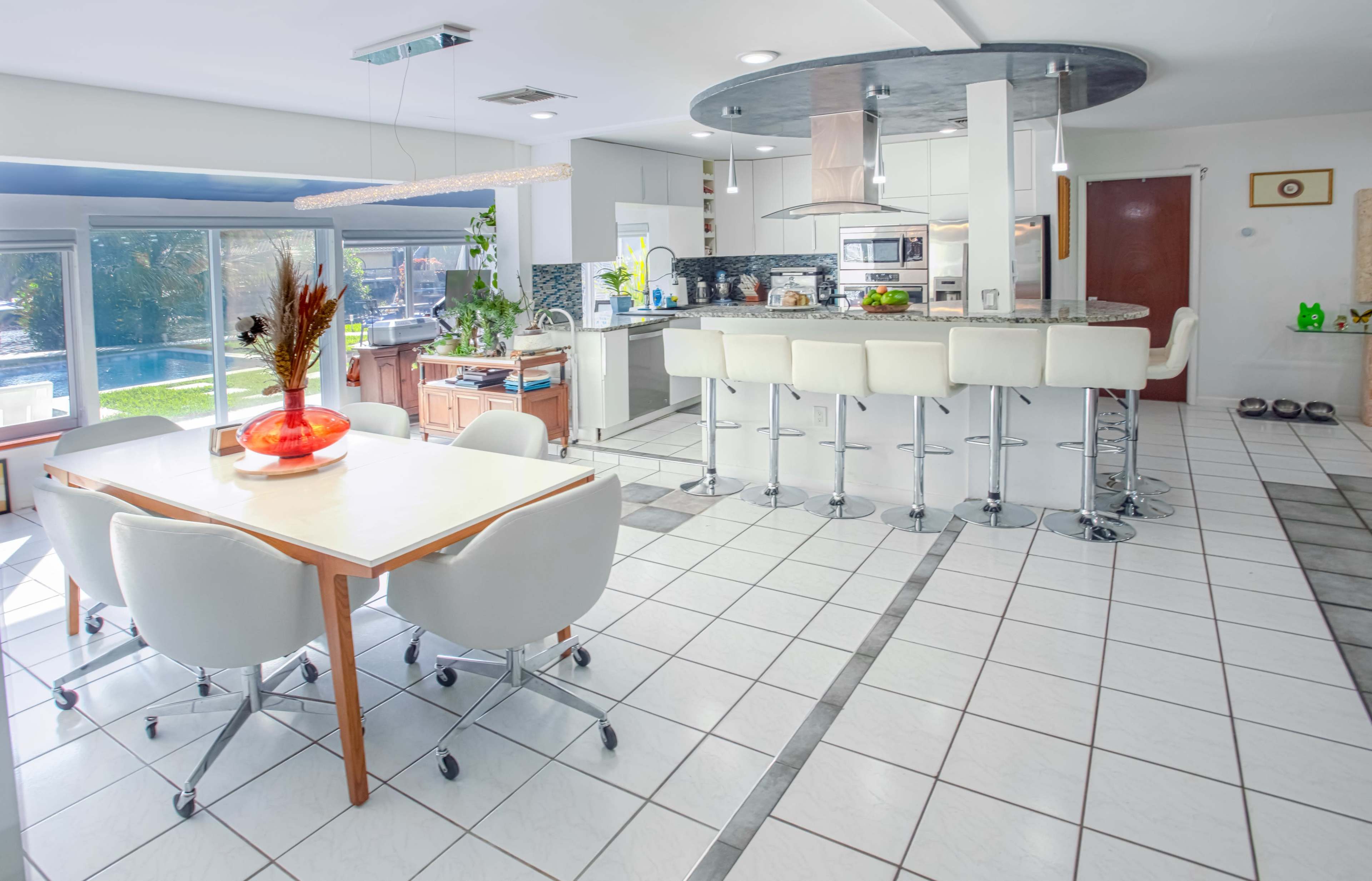 A modern kitchen and dining area features a white table surrounded by rolling chairs, with a bar counter and stools, large windows, and a view of a pool outside.