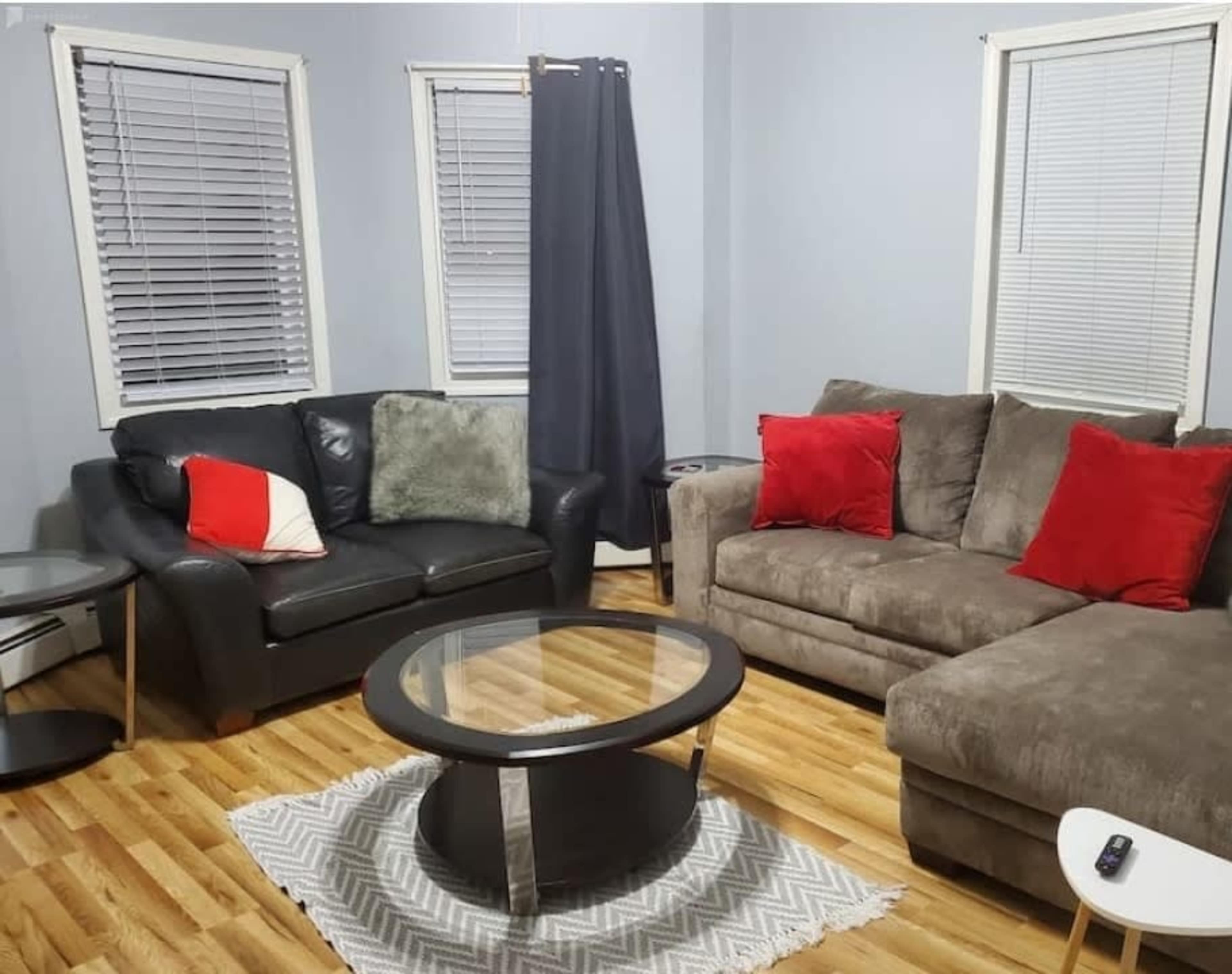 A living room features a black leather couch, a gray sectional sofa with red throw pillows, a round glass coffee table, and wooden floors.