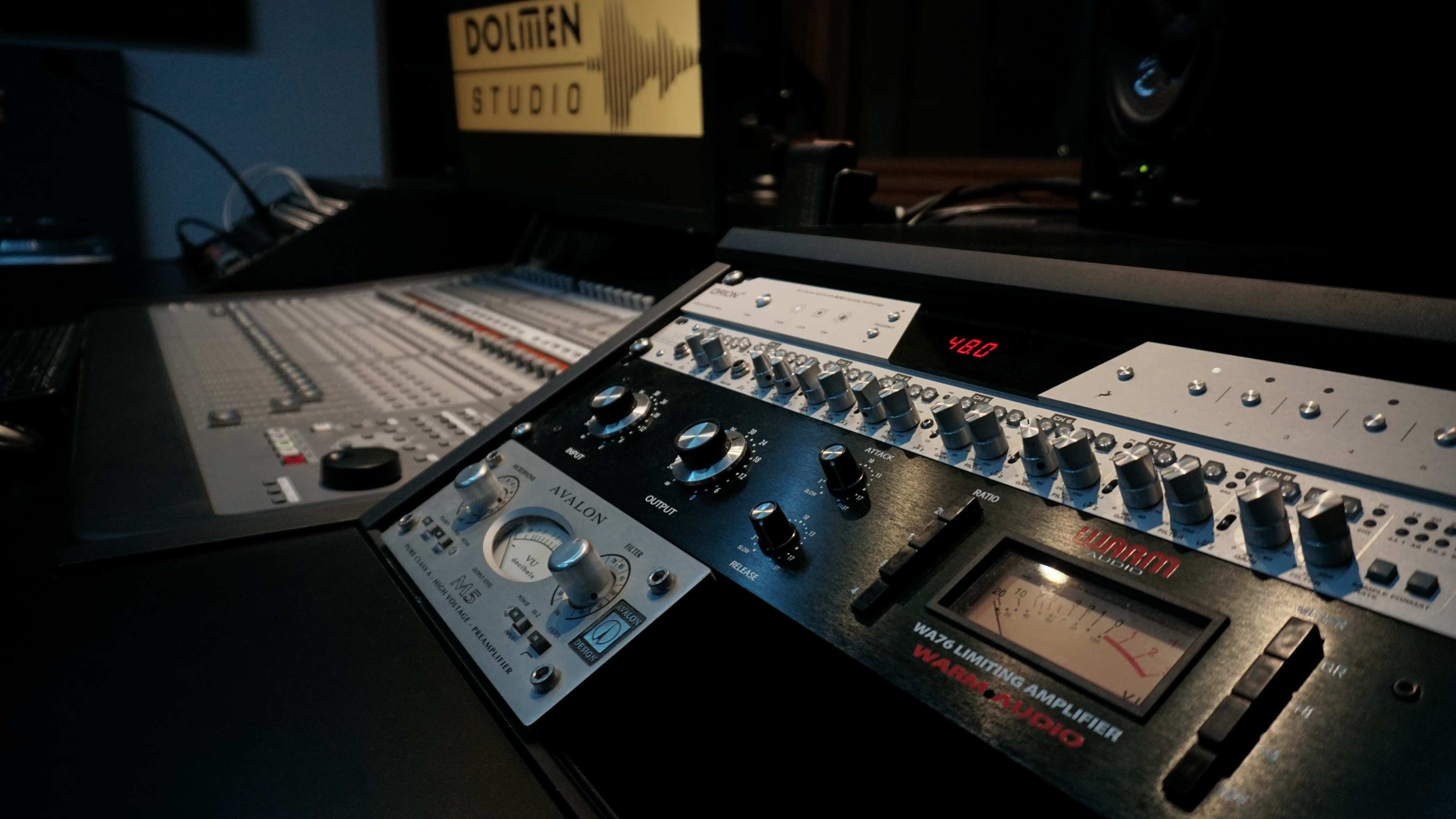 The image shows a close-up of a sound mixing console with various knobs and meters, set against a blurred studio backdrop featuring a sign.