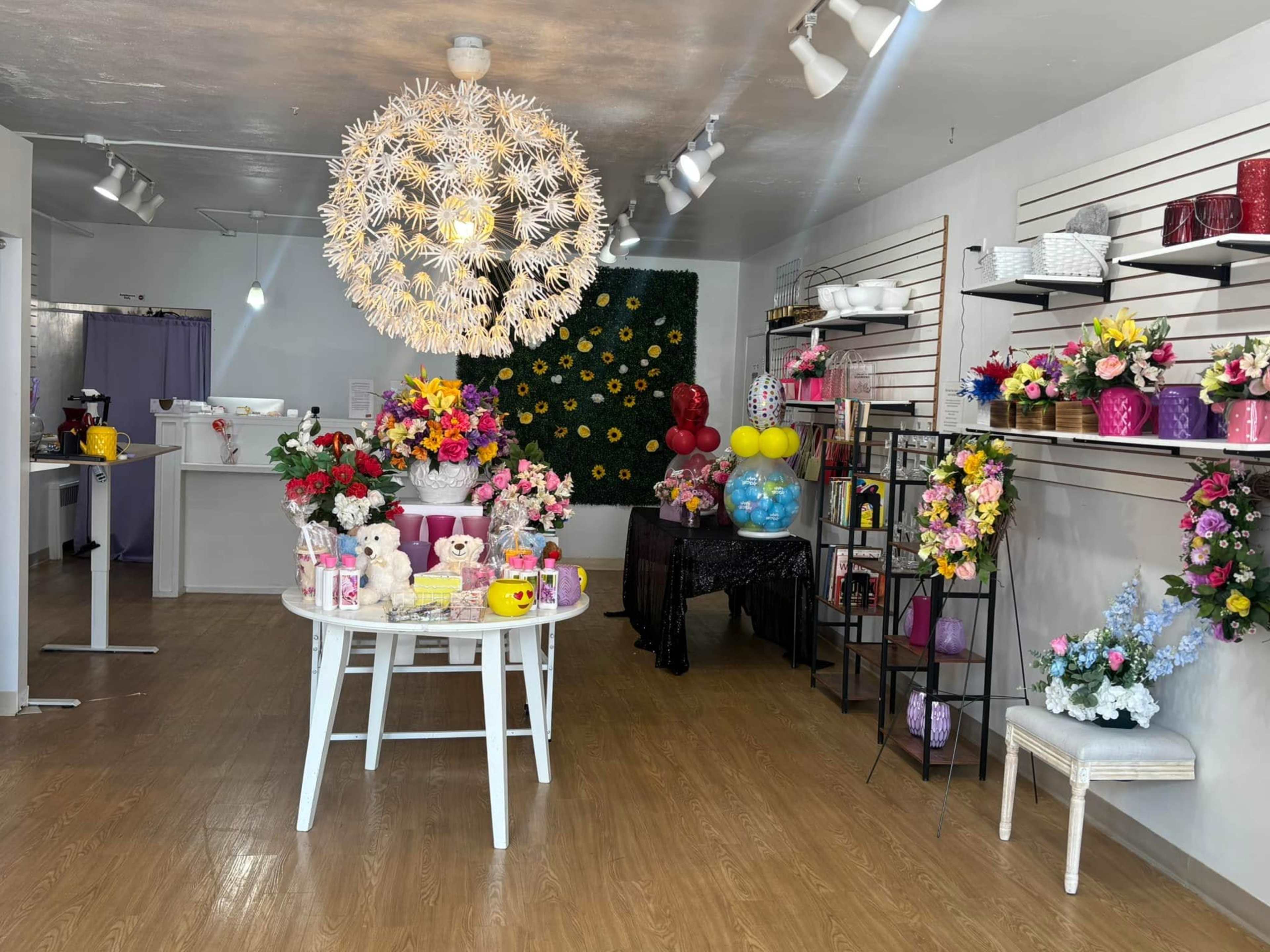 The image shows a floral shop with various colorful arrangements displayed on shelves and a central table adorned with flowers, teddy bears, and balloons.