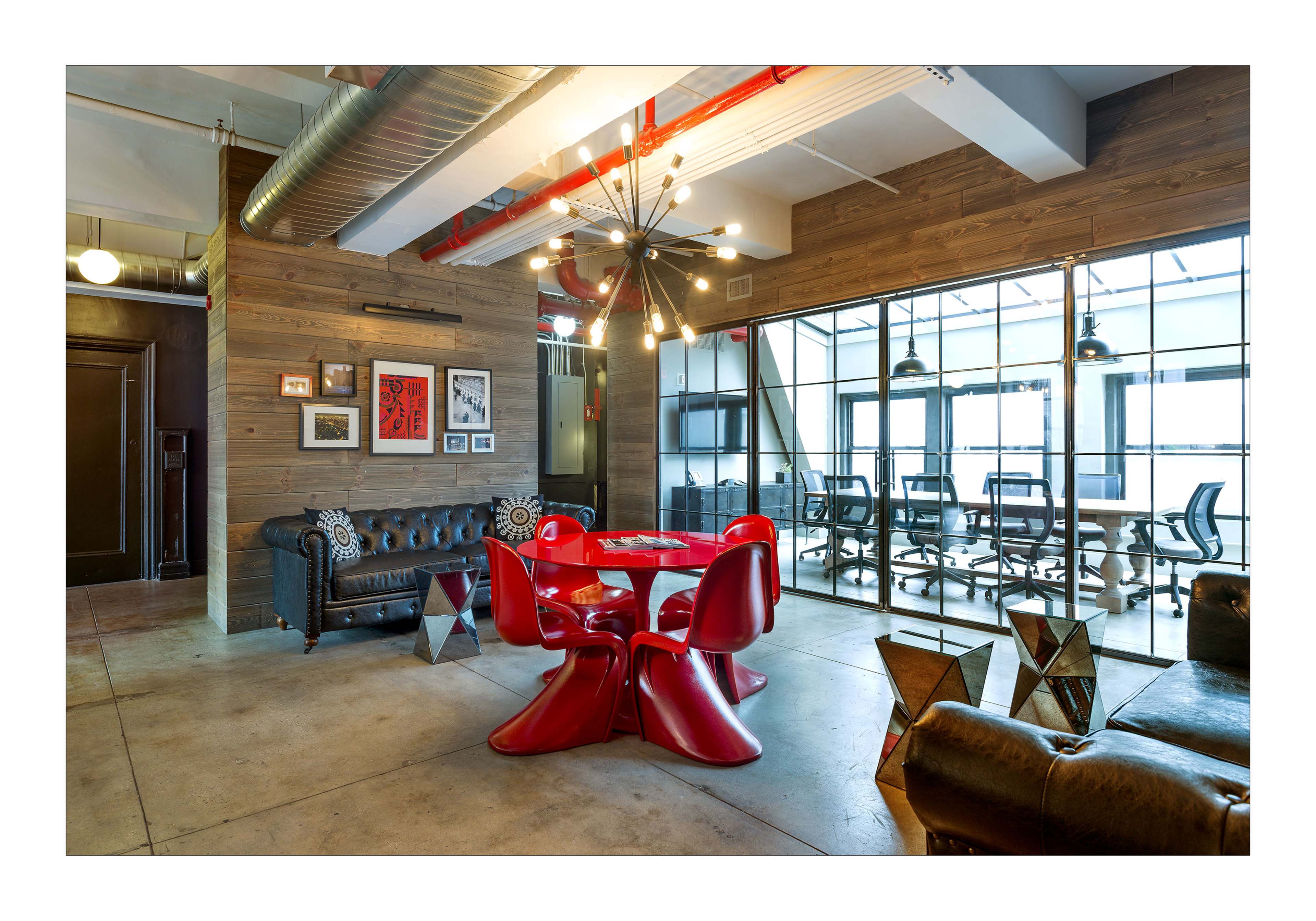 A modern office space featuring a red dining table surrounded by matching chairs, a black leather couch, and large windows with a conference room visible in the background.