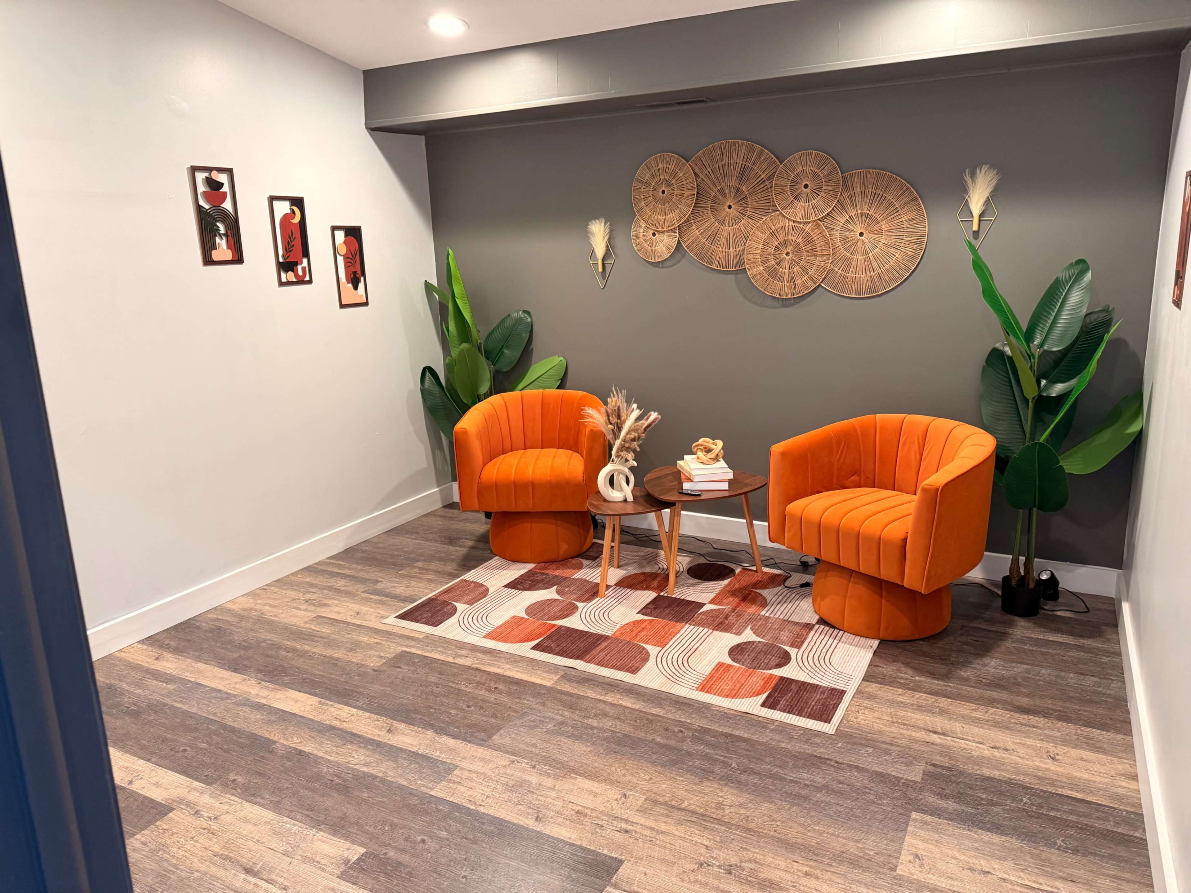 The image shows a cozy interior space featuring two orange armchairs, a small round table, and decorative wall art, with plants adding greenery to the room.