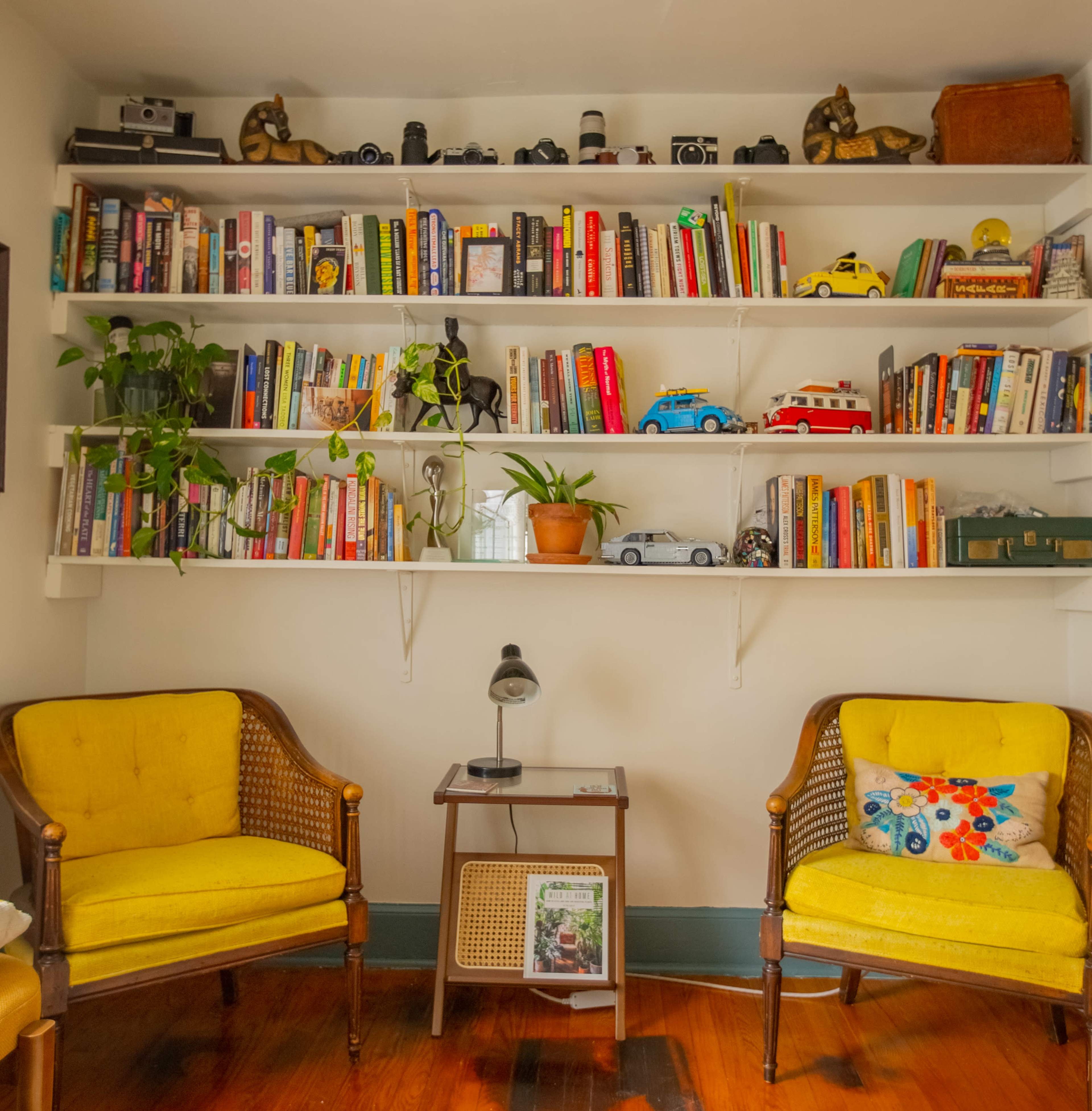 The image shows a cozy reading nook with two yellow chairs facing a small table, surrounded by a shelf filled with books, plants, and decorative items.