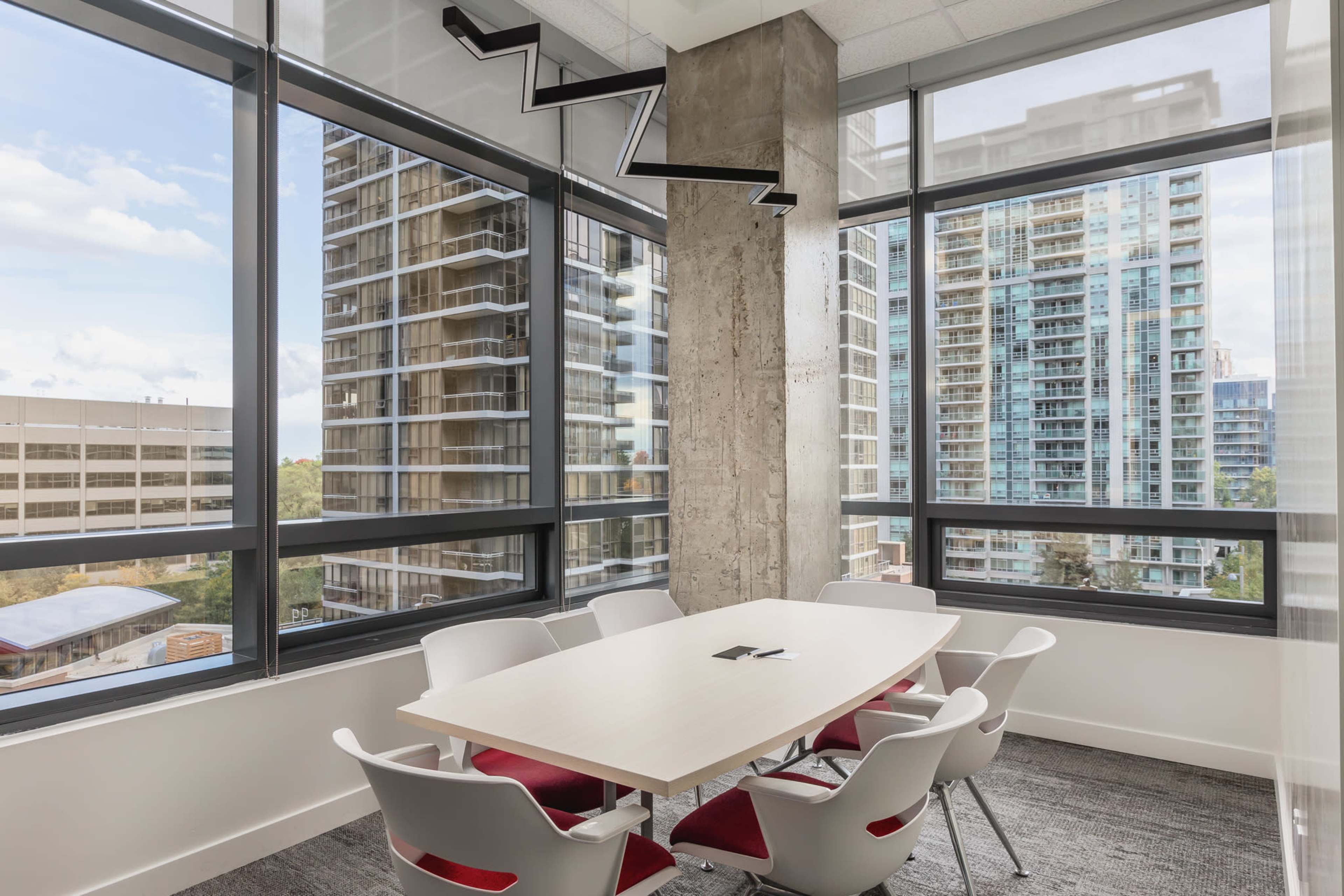 A conference room with a large table surrounded by chairs, featuring floor-to-ceiling windows that showcase urban buildings.