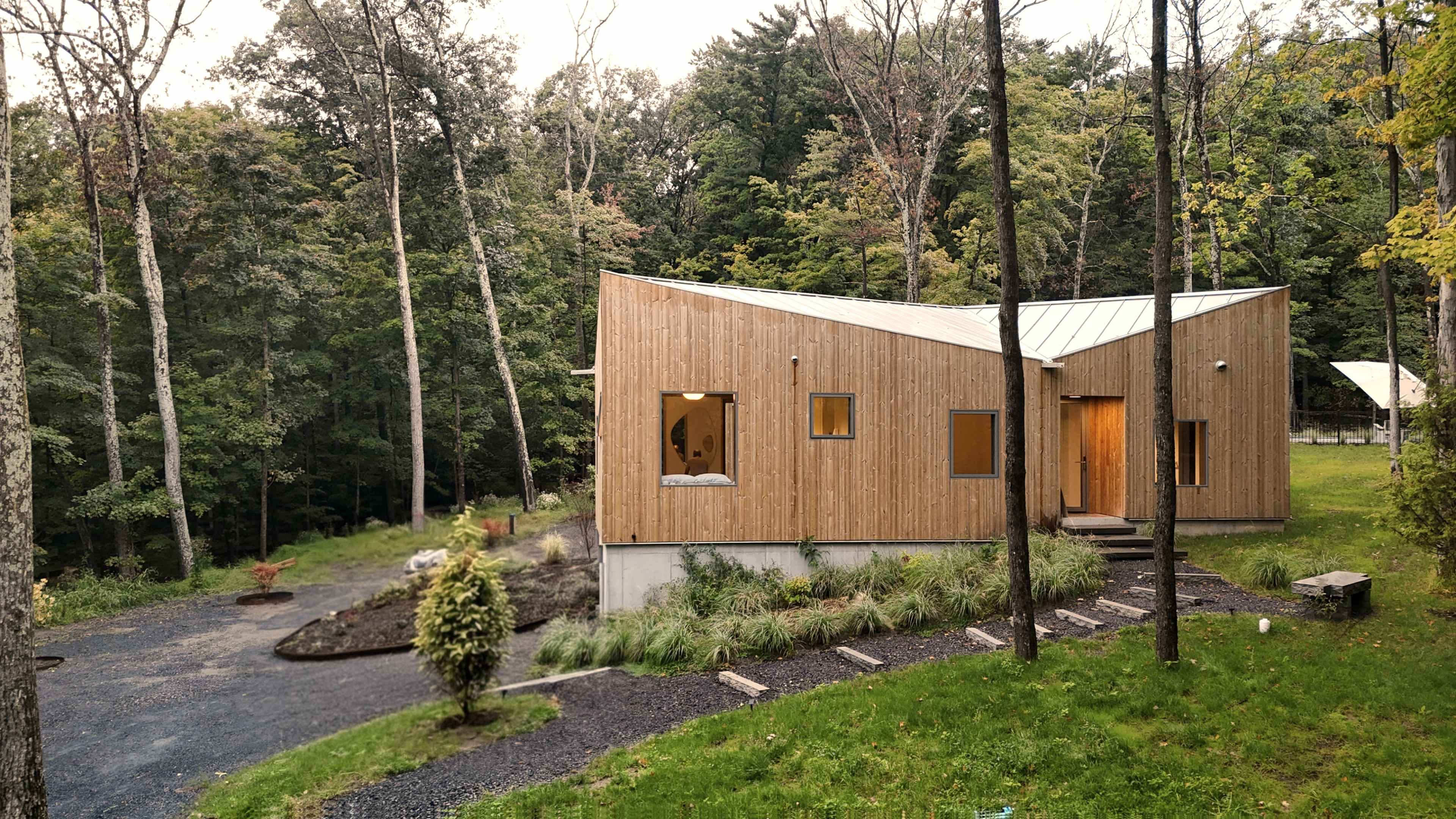 A modern wooden house with large windows sits among trees, featuring a gravel driveway and landscaped grass areas.