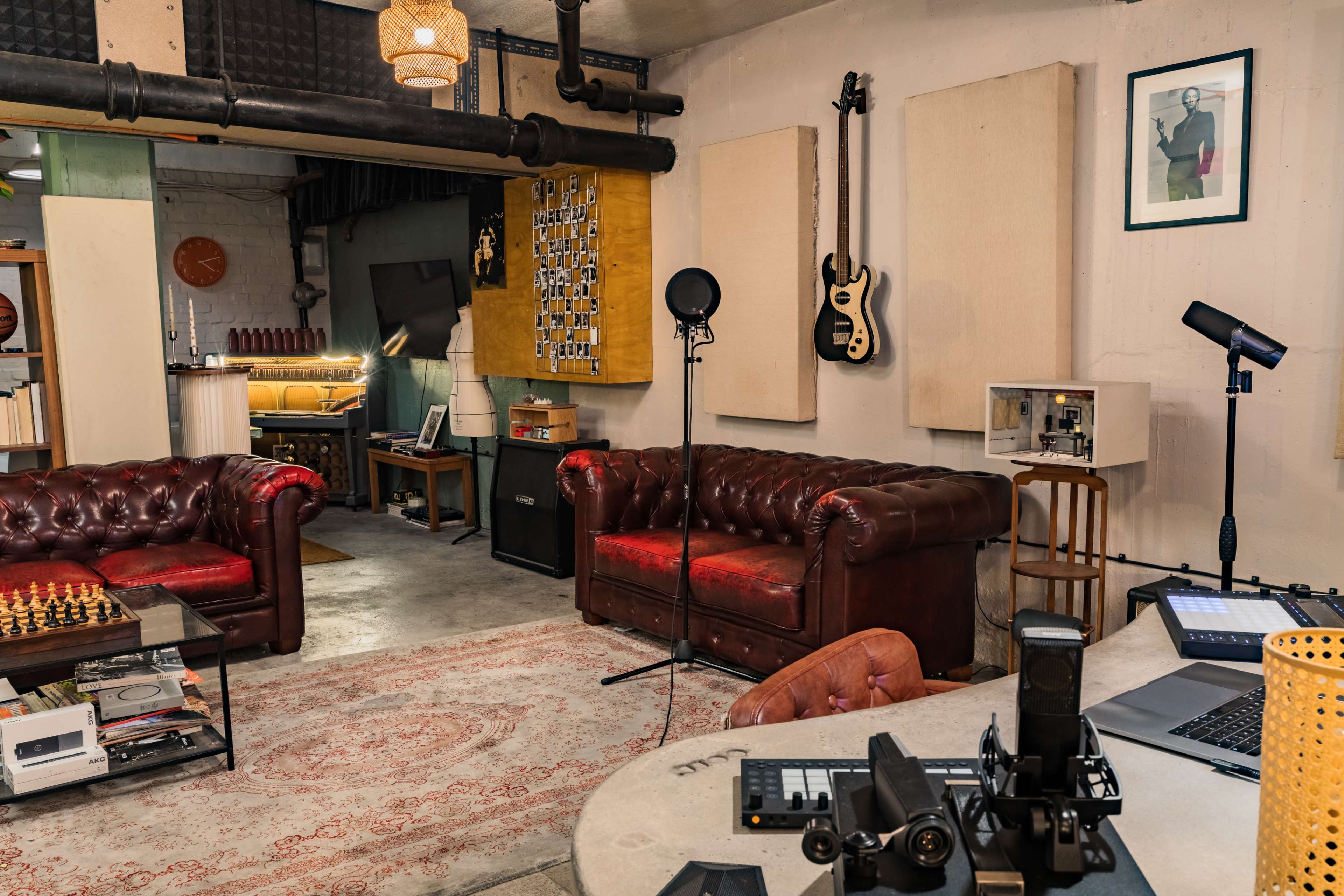 A modern studio space features two brown leather sofas, a coffee table with a chess set, musical instruments on the wall, and recording equipment on a desk.