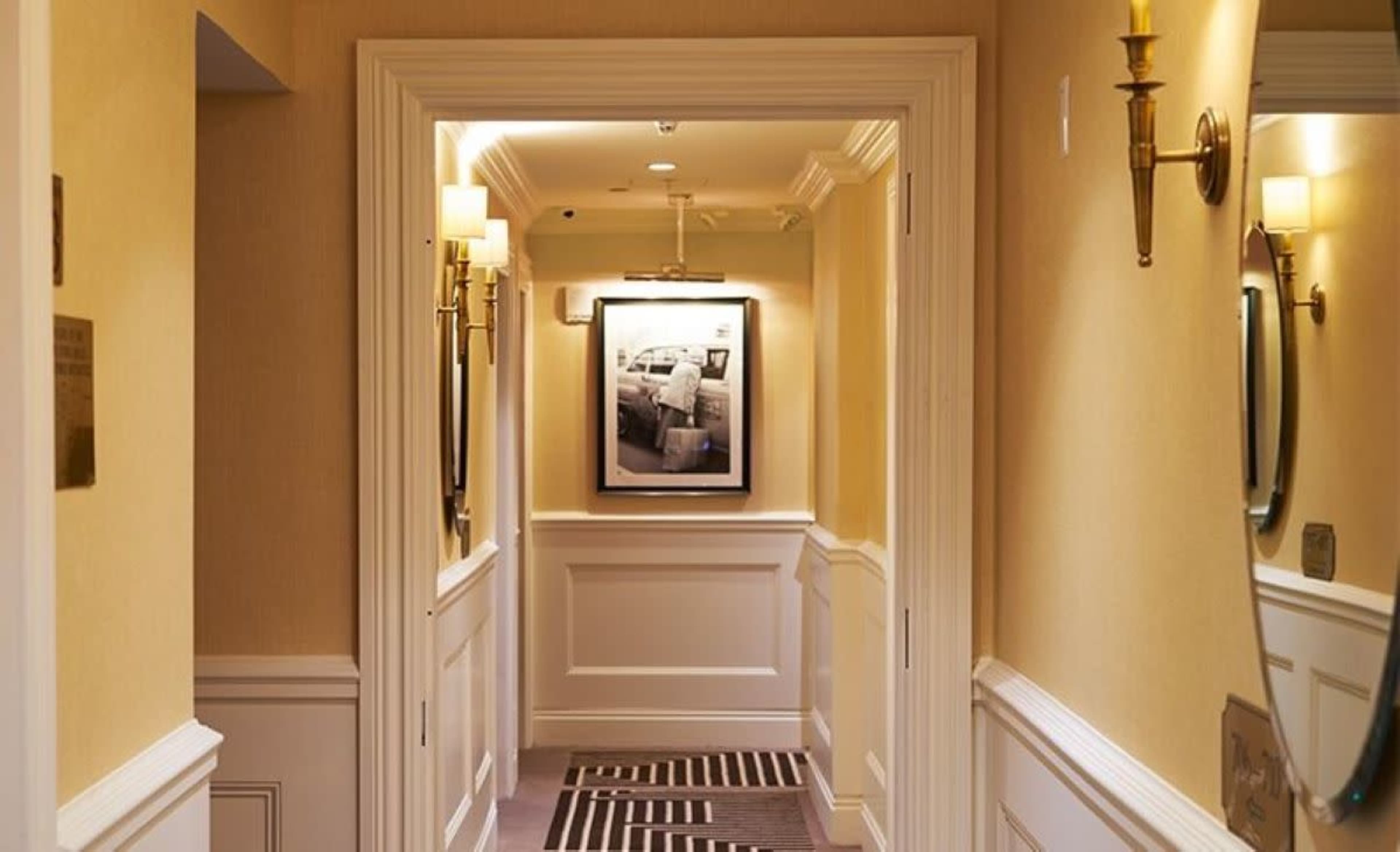 A narrow corridor features yellow walls, wall sconces, a large framed photograph, and a floor rug with a striped pattern.