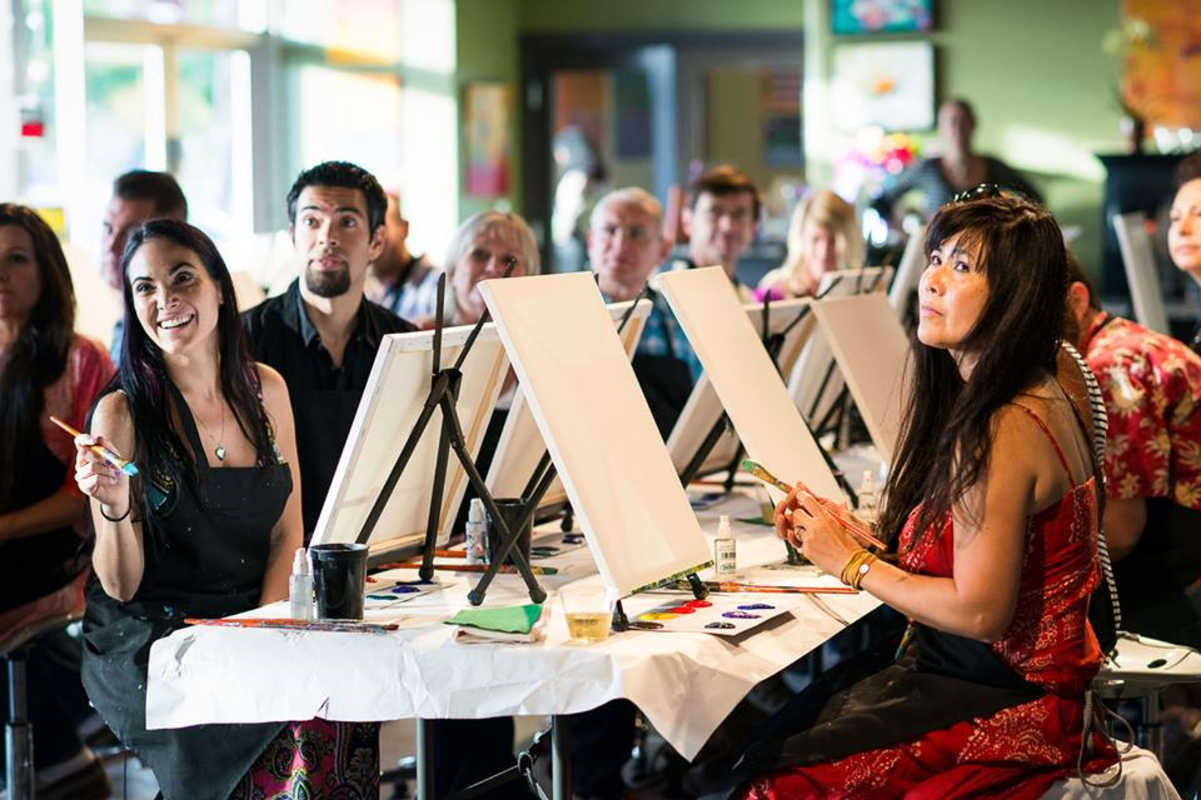 A group of people sit at tables in an art class, facing blank canvases as two women hold paintbrushes.