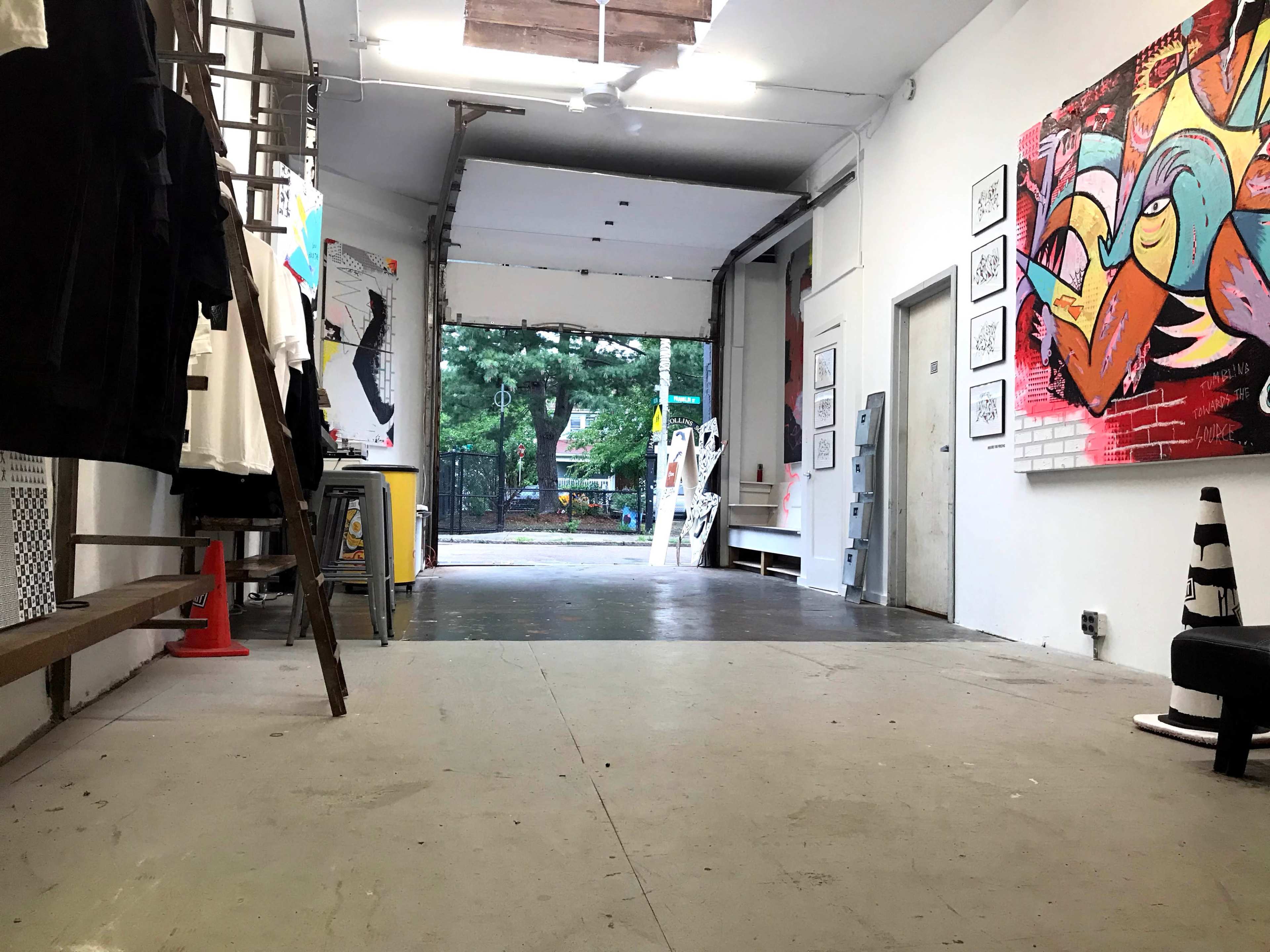 An industrial-style gallery space with an open garage door leading to a green outdoor area, featuring artworks on the walls and various items arranged inside.