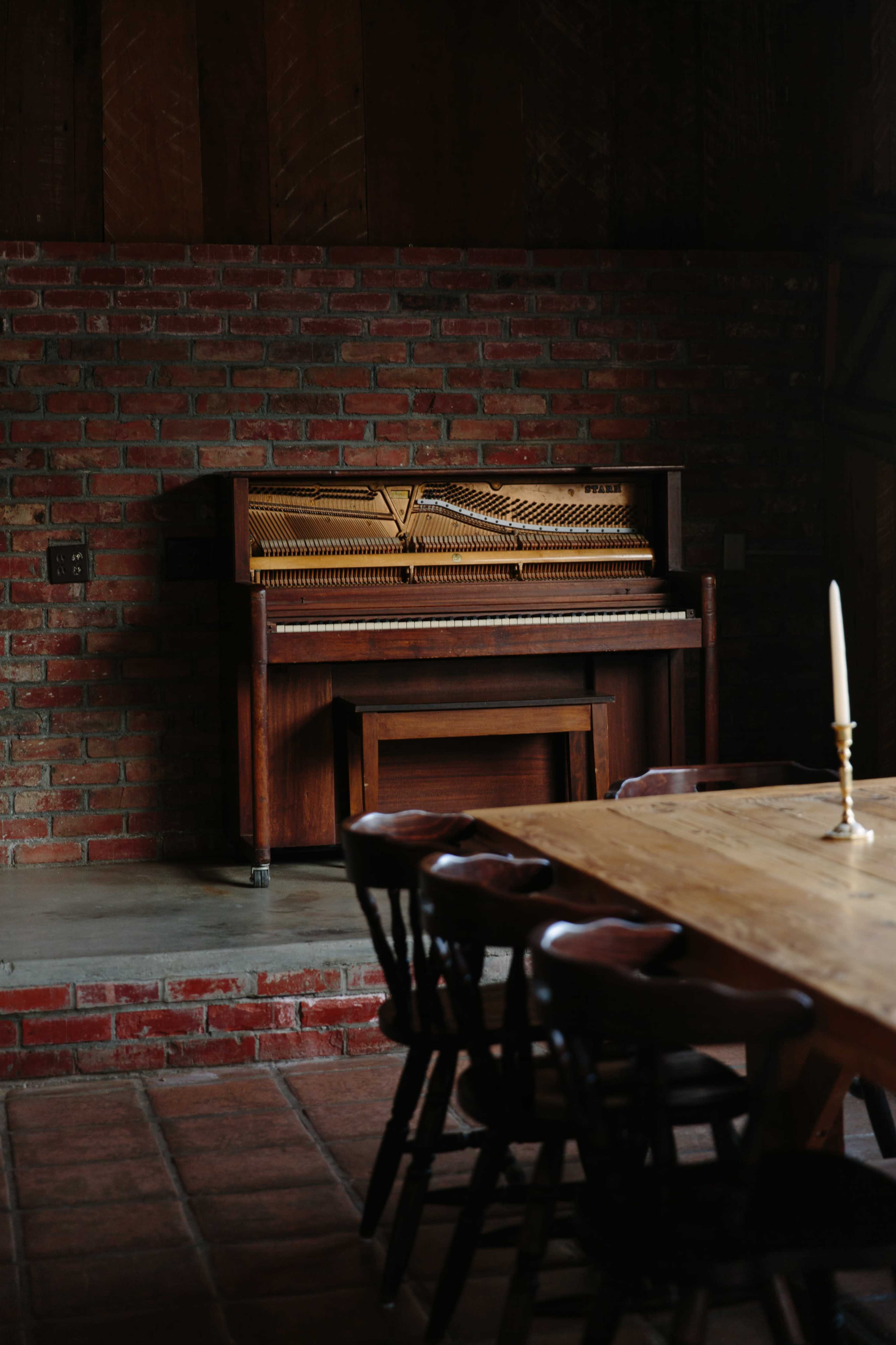 A wooden piano sits against a brick wall next to a long wooden dining table with chairs.