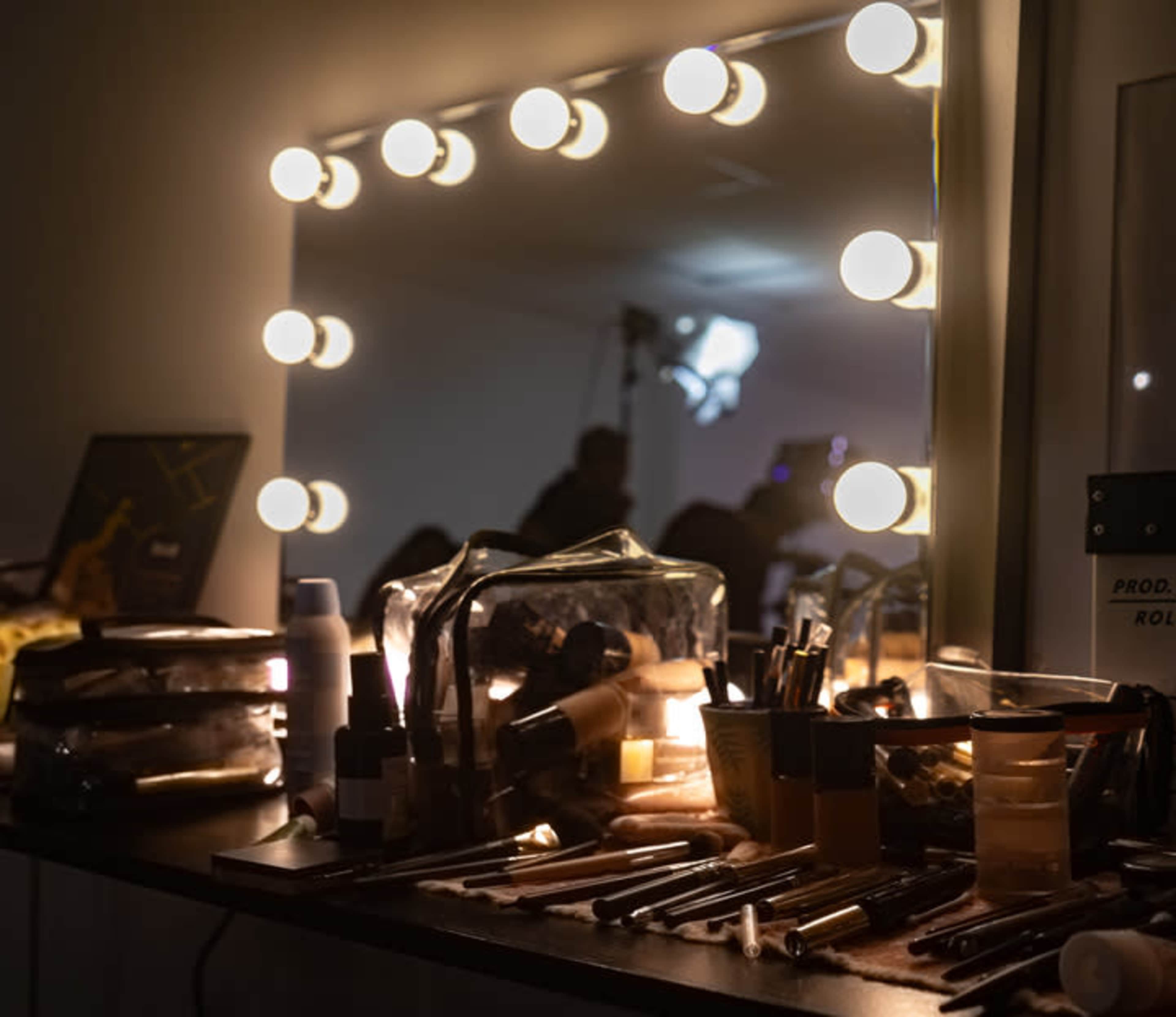 A makeup station is illuminated by bright bulbs, with various cosmetics and brushes arranged on the counter.
