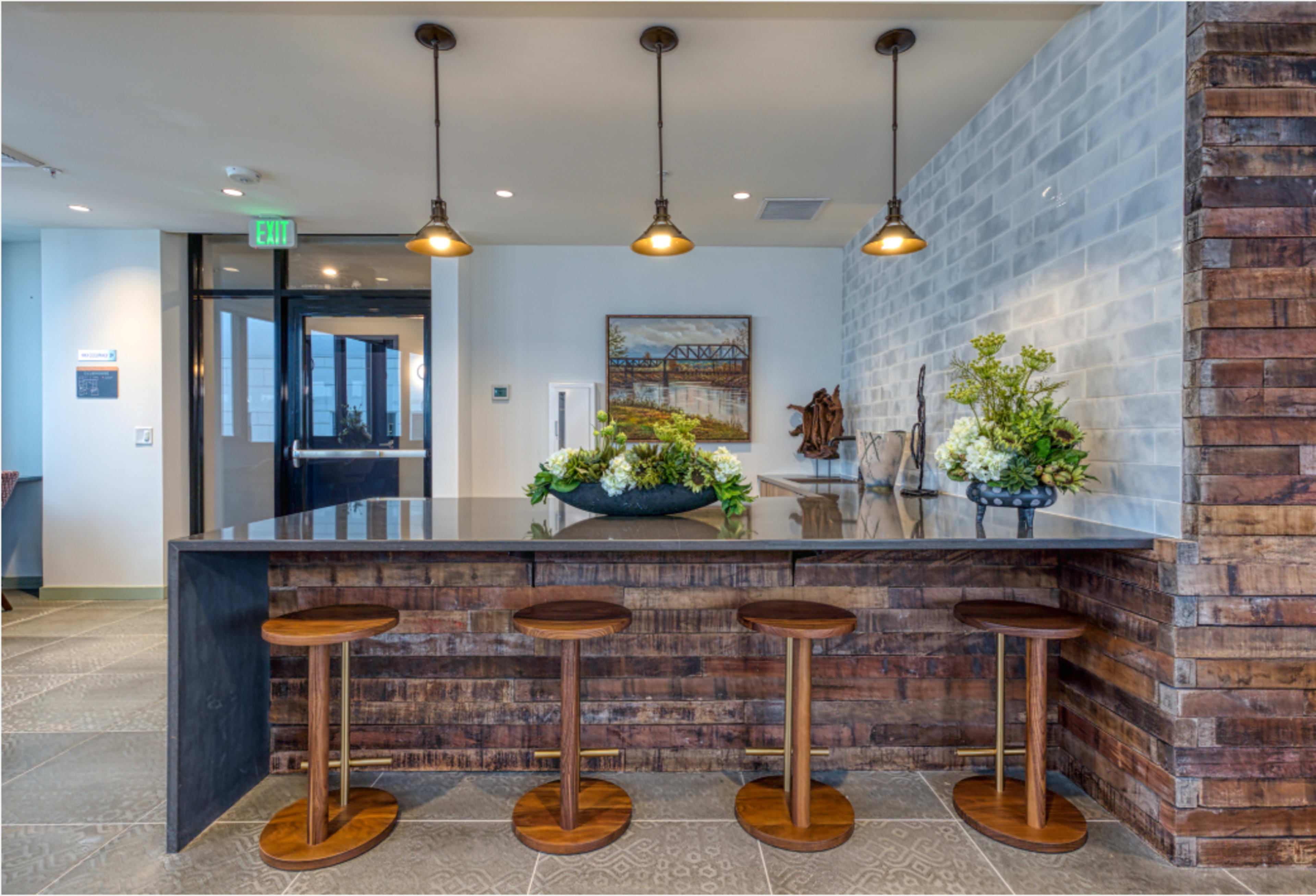 The image shows a modern bar area with a dark countertop, wooden stools, and decorative plants against a textured wall.