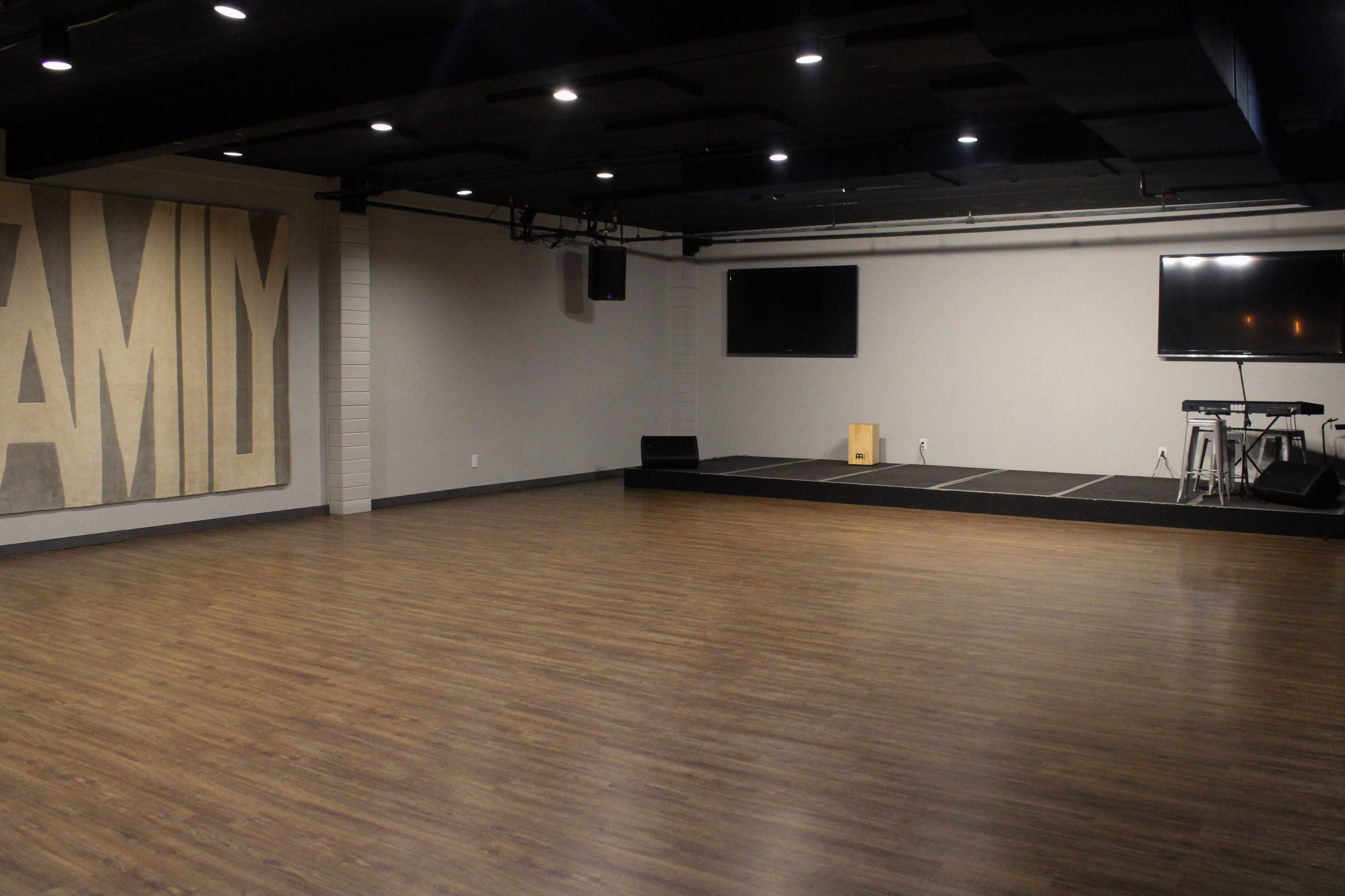 An empty indoor space with a wooden floor, a large wall mural that reads "FAMILY," and stage equipment including monitors and a keyboard setup.