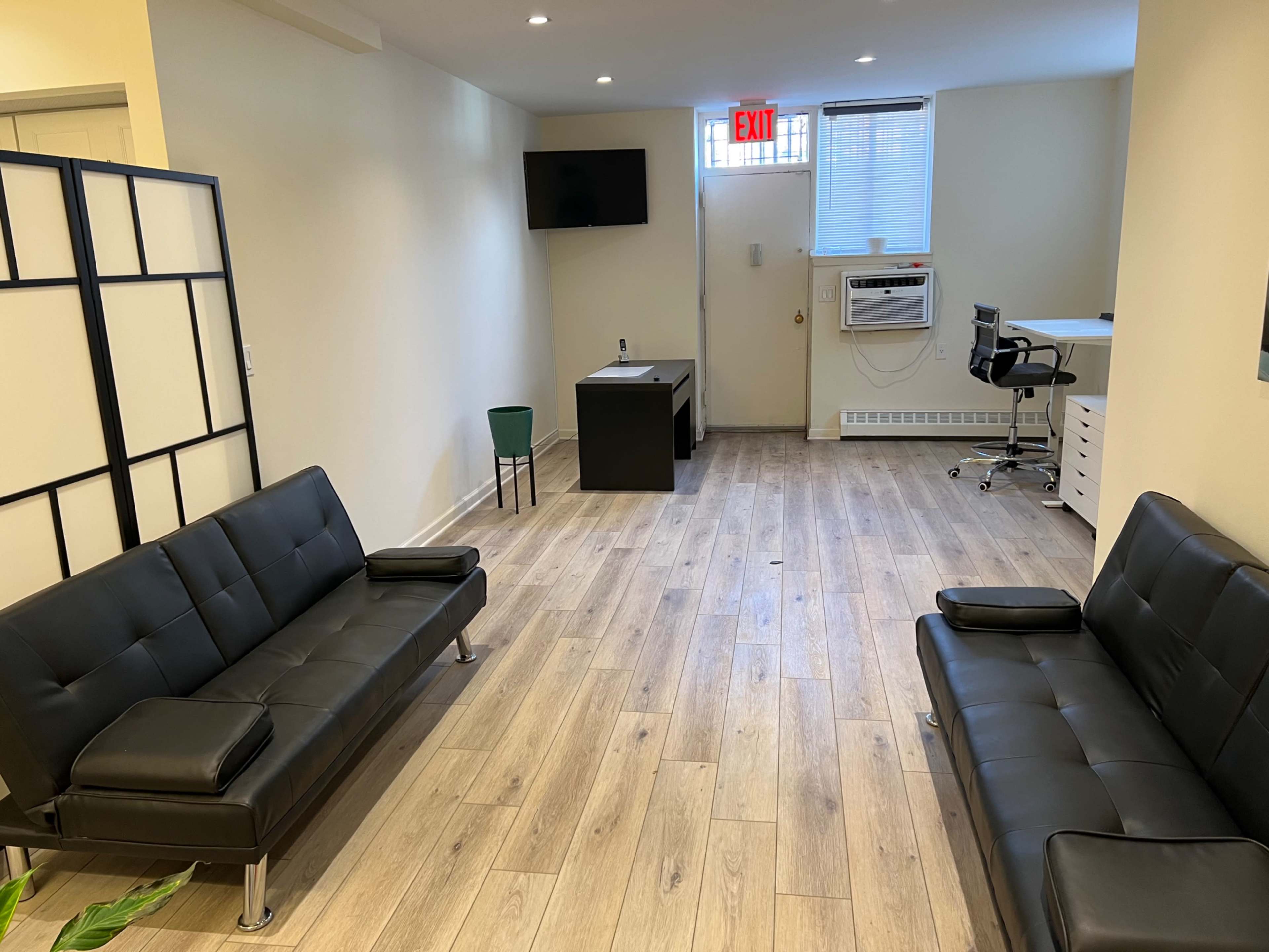 A light-colored room with wooden flooring, featuring two black sofas, a small green chair, a desk with a chair, and an air conditioning unit on the wall.