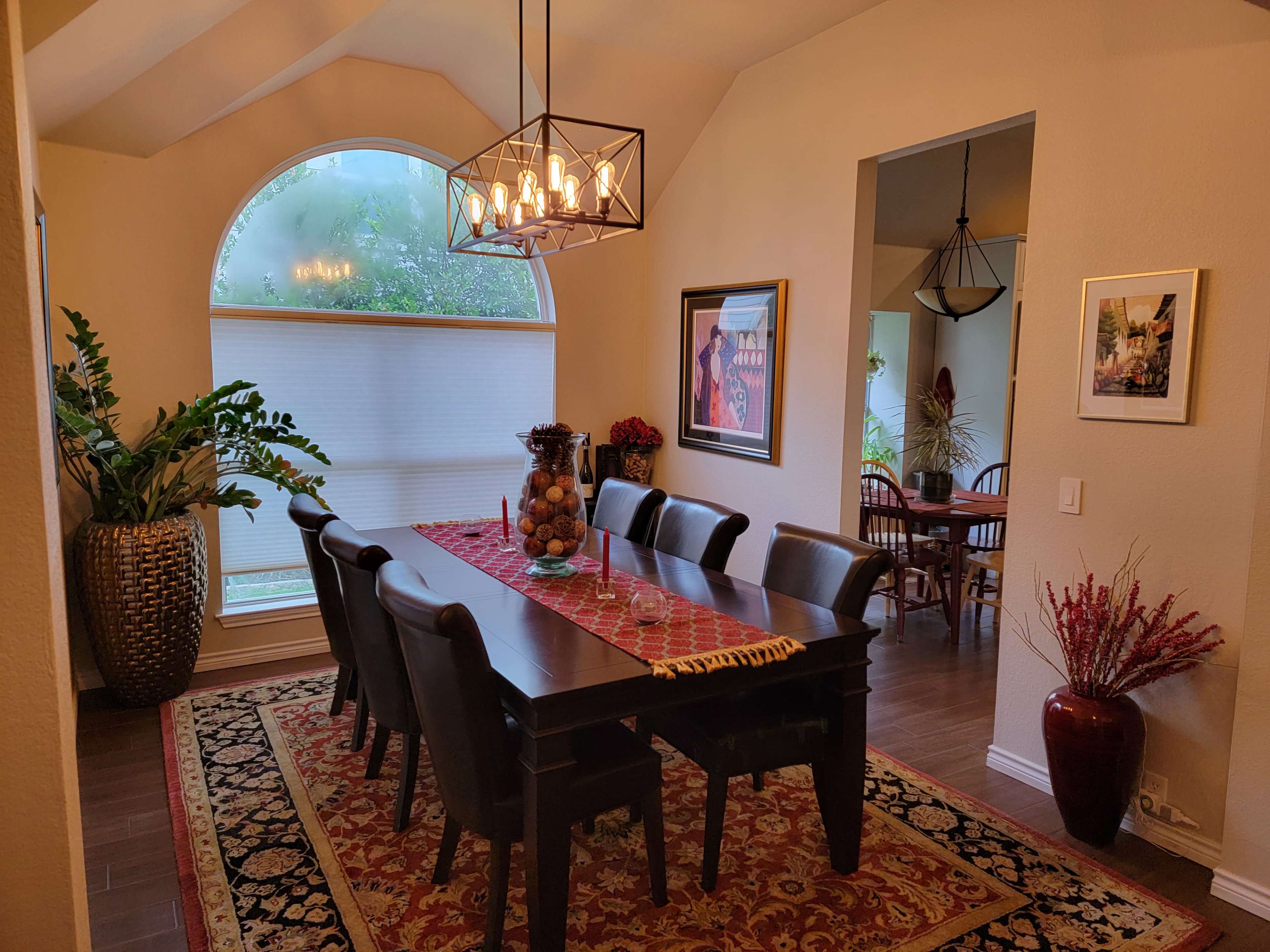 A spacious dining area features a long wooden table surrounded by eight chairs, with a large window and decorative plants in the background.