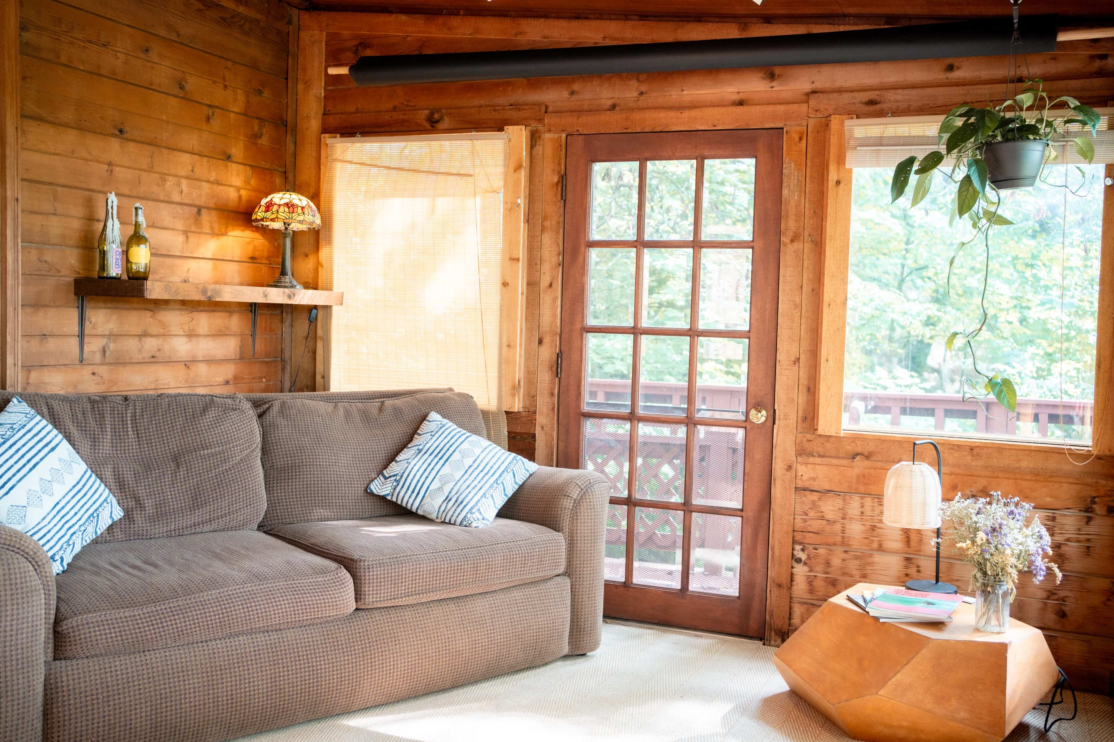 The image shows a cozy wooden interior featuring a sofa with decorative cushions, a glass door leading to the outside, and a small table with a vase of flowers.