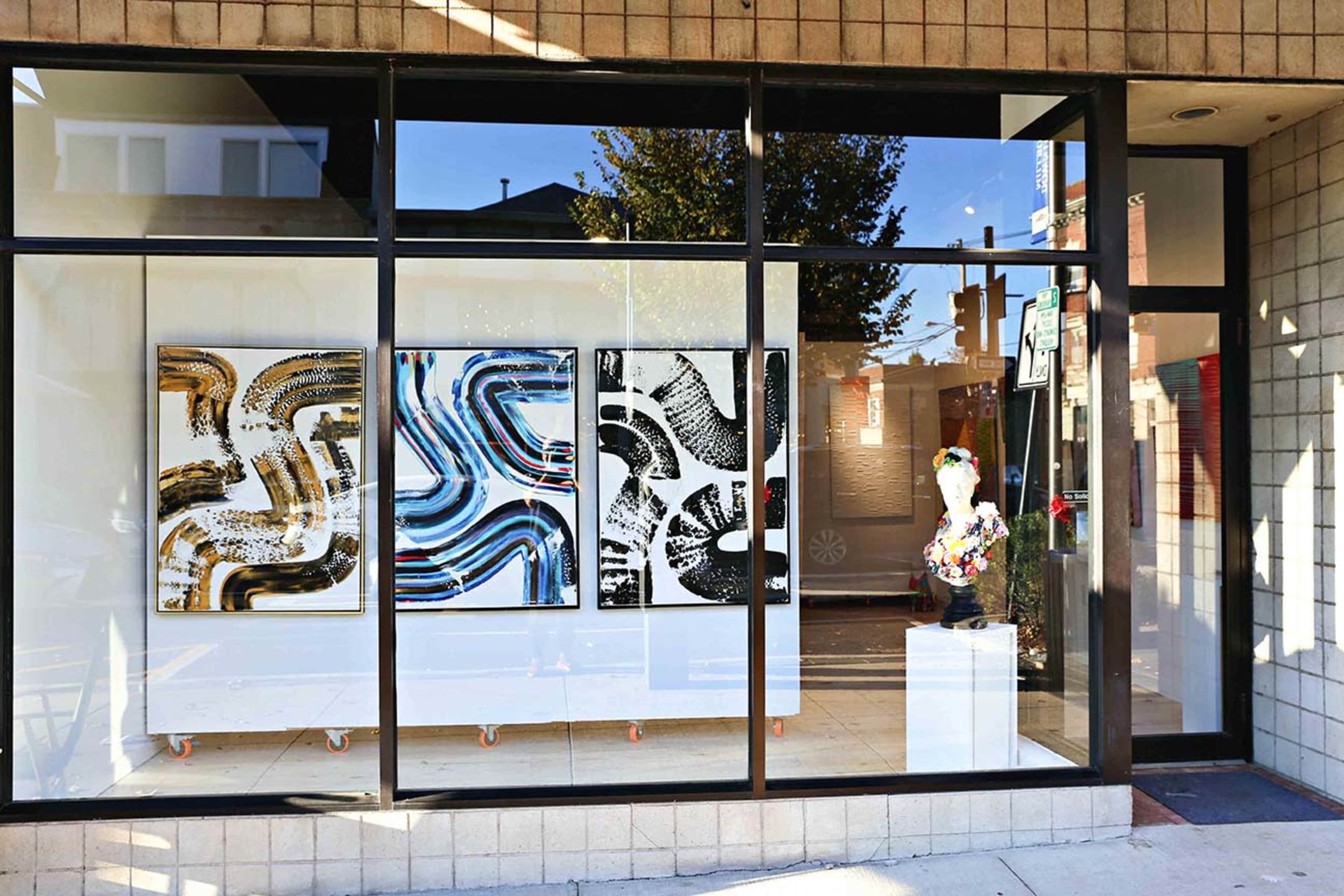 A modern art gallery window displaying three abstract paintings and a sculptural piece on a white pedestal.