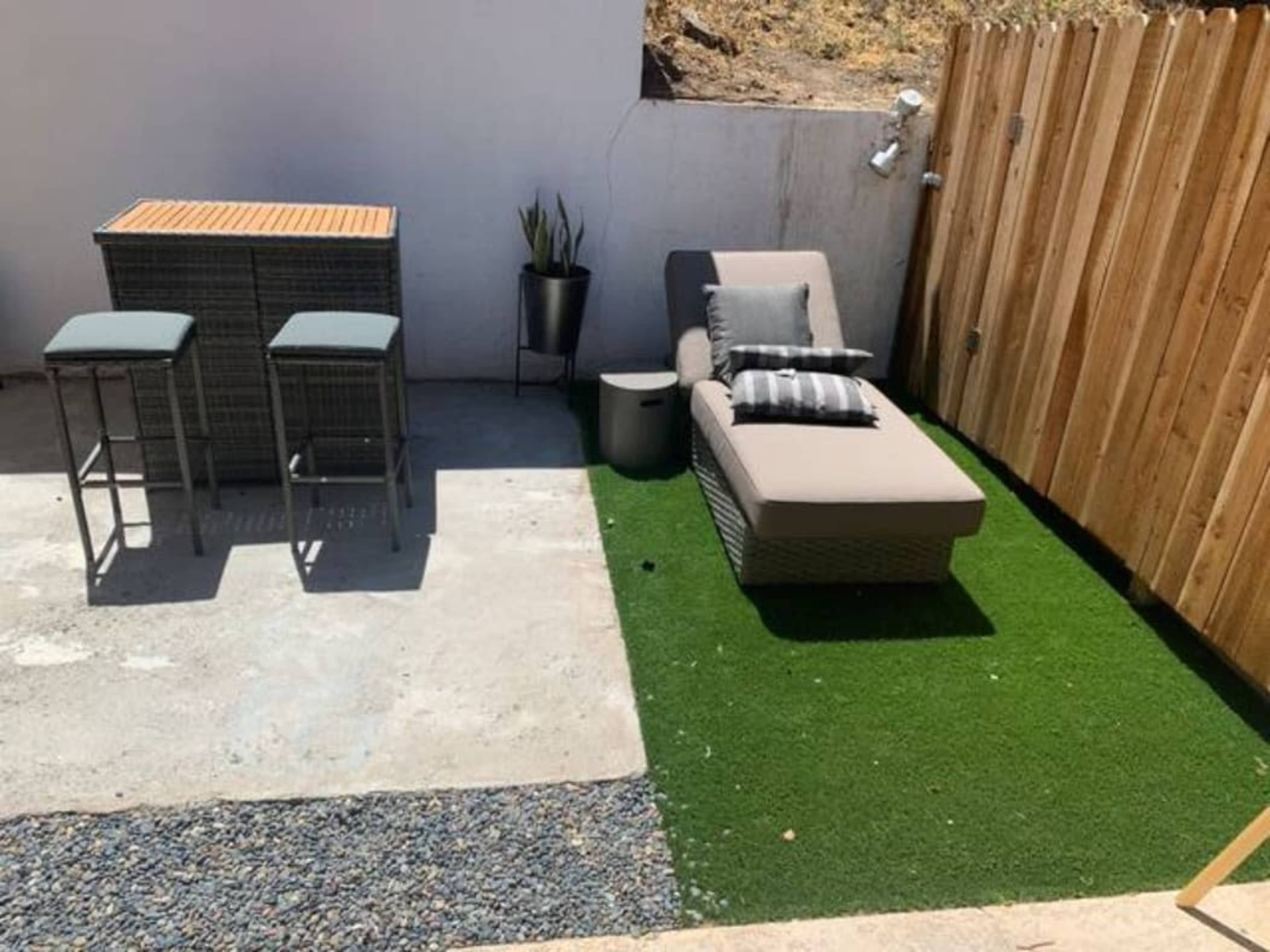 A compact outdoor space features a lounge chair with cushions, a small side table, a bar with two stools, and artificial grass bordered by gravel and wooden fencing.