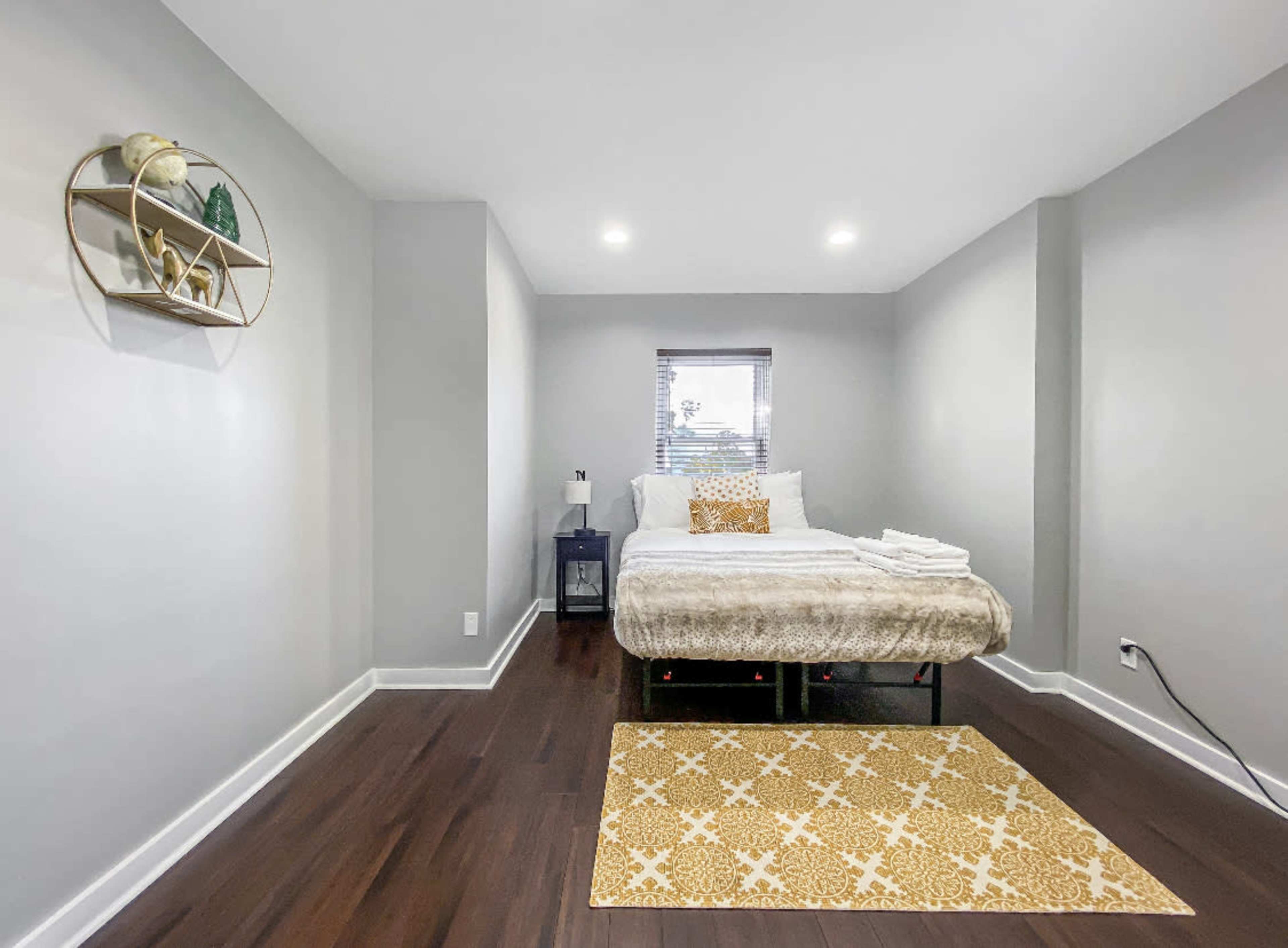 A neatly arranged bedroom features a bed with a light-colored blanket, a nightstand, and a decorative wall shelf, all set against gray walls and dark wooden flooring.