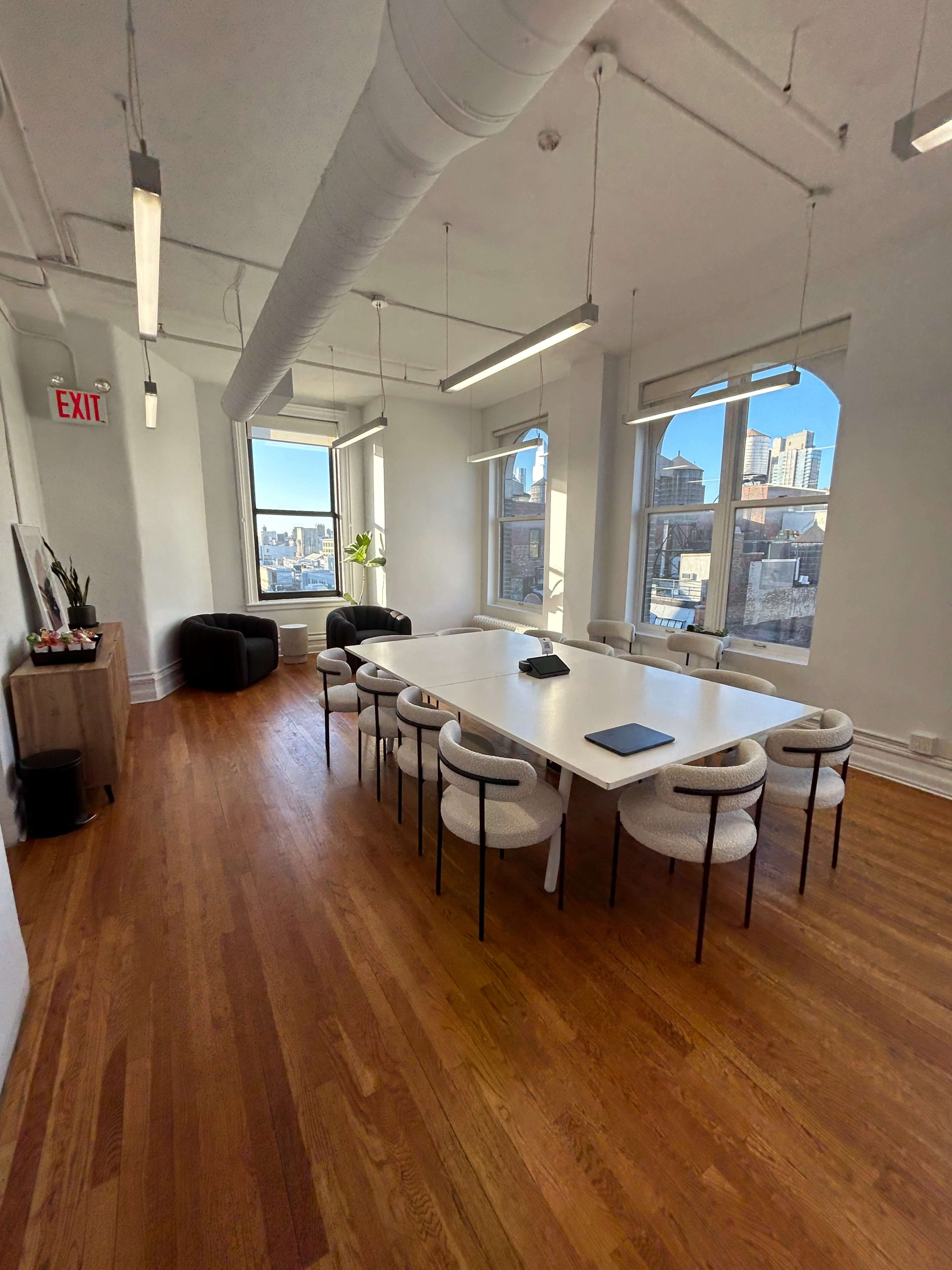 A modern conference room features a long white table surrounded by upholstered chairs, with large windows showcasing a city skyline.