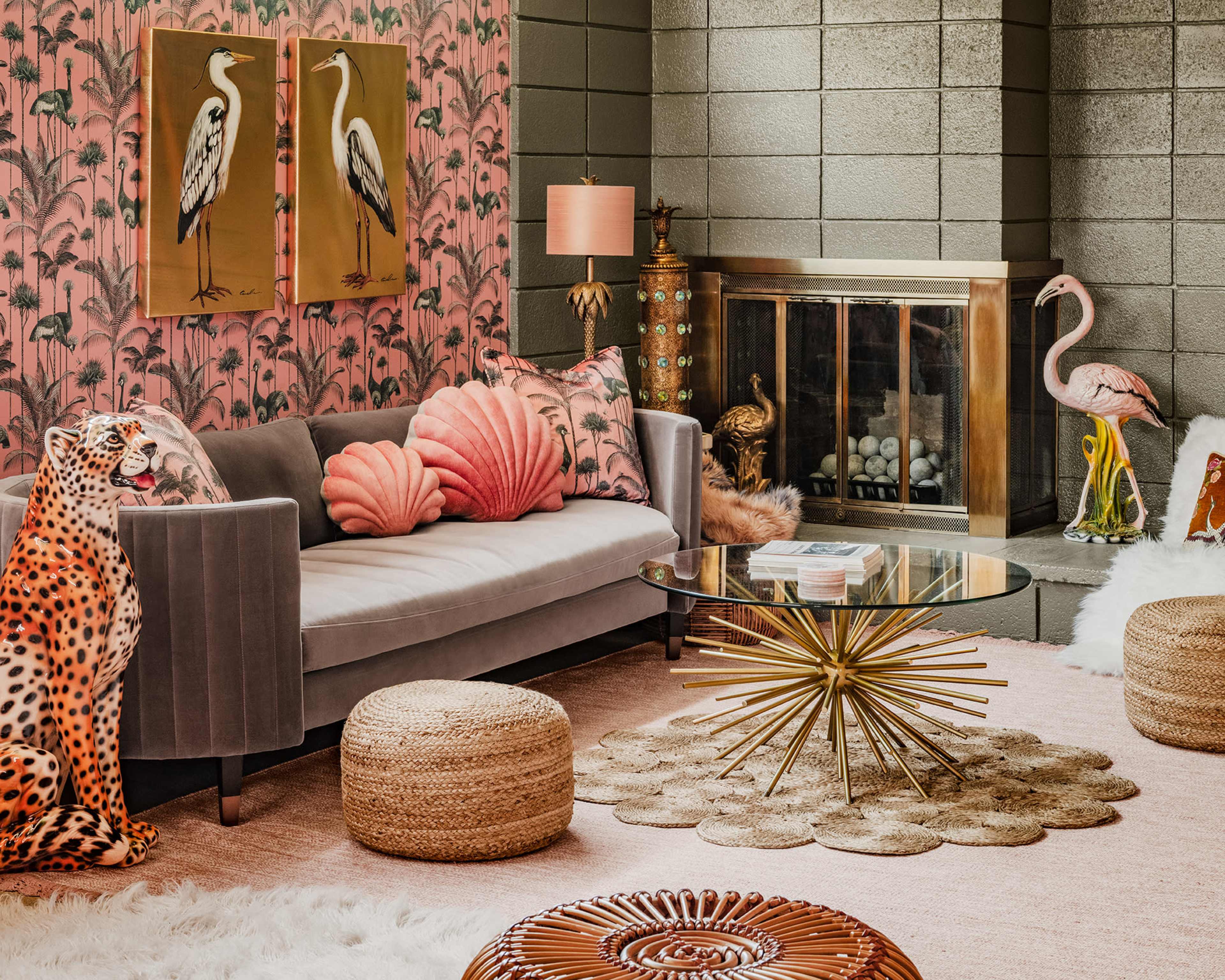 The living room features a gray sofa adorned with decorative pillows, a round glass coffee table, and various artistic elements, including tropical-themed wallpaper and a fireplace.