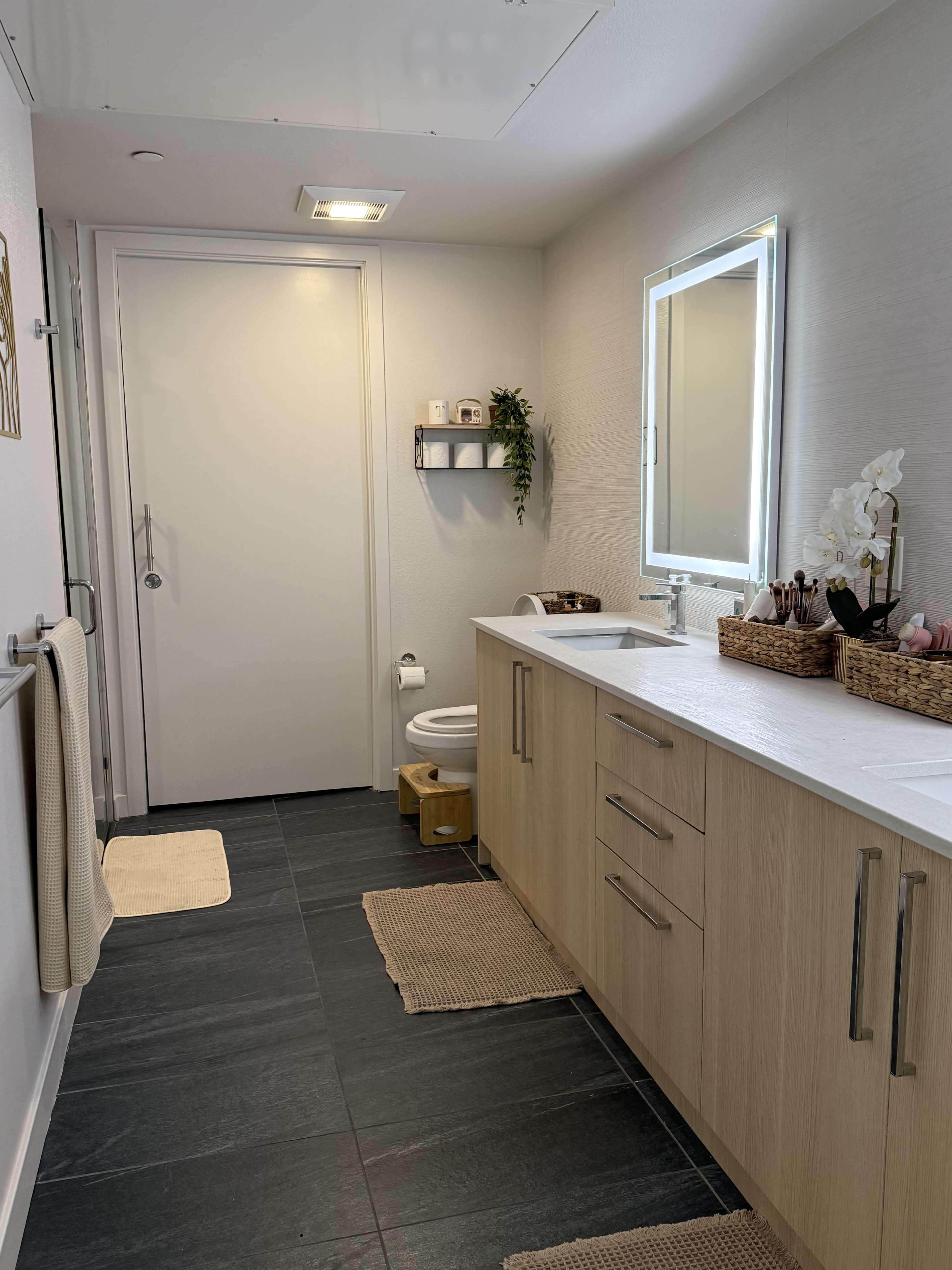 The image shows a modern bathroom with a wooden vanity, a large mirror, and dark tile flooring.
