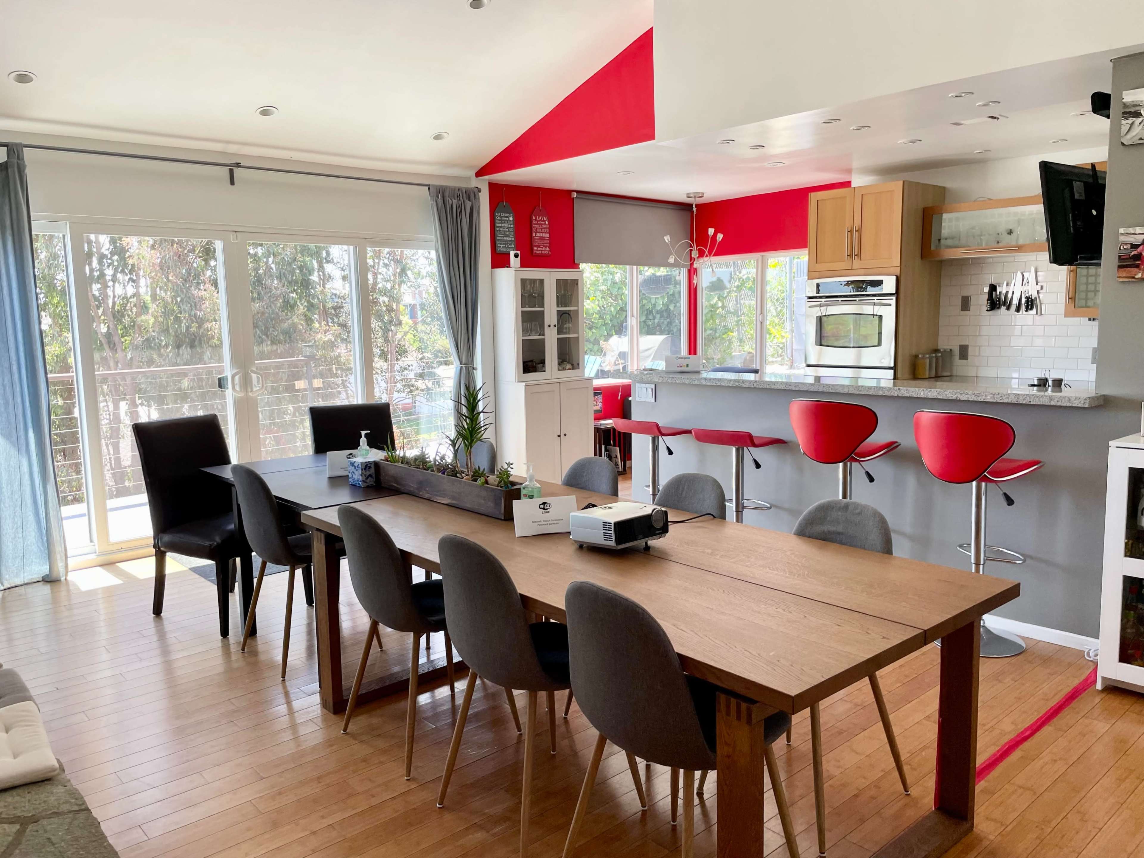 A modern kitchen and dining area features a long wooden table with gray chairs, red bar stools at a countertop, and large windows offering views of the outdoors.