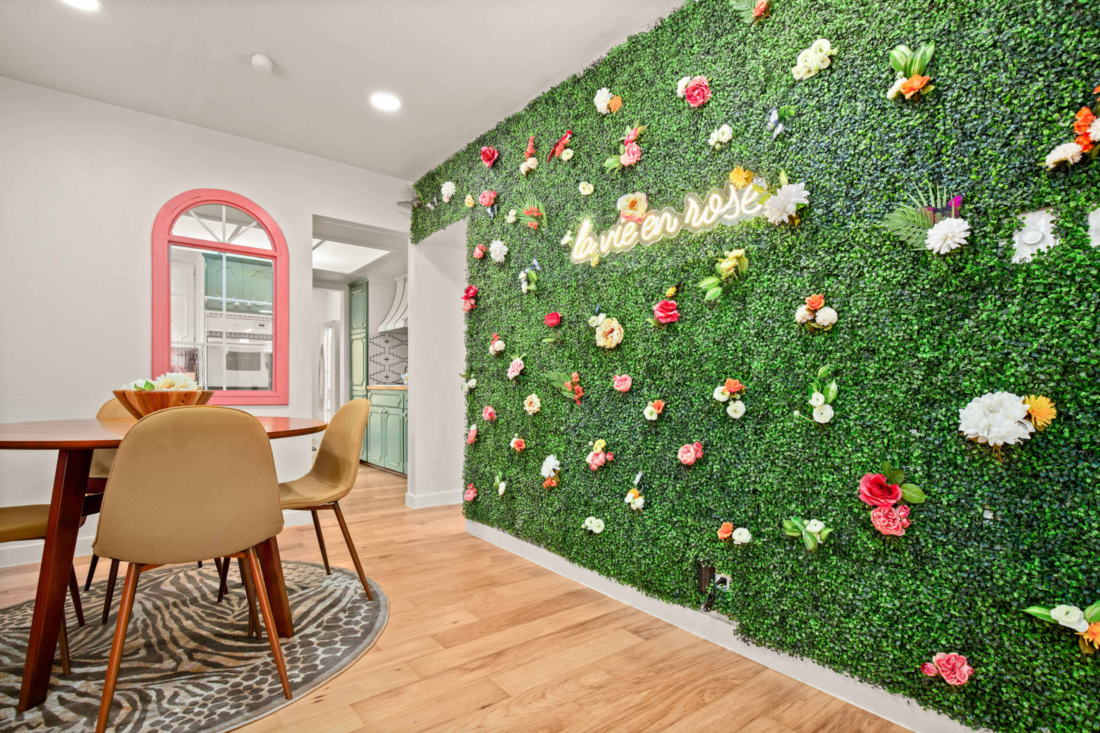 The image shows a dining area with a wooden table surrounded by chairs, next to a decorative wall covered in faux greenery and colorful artificial flowers.