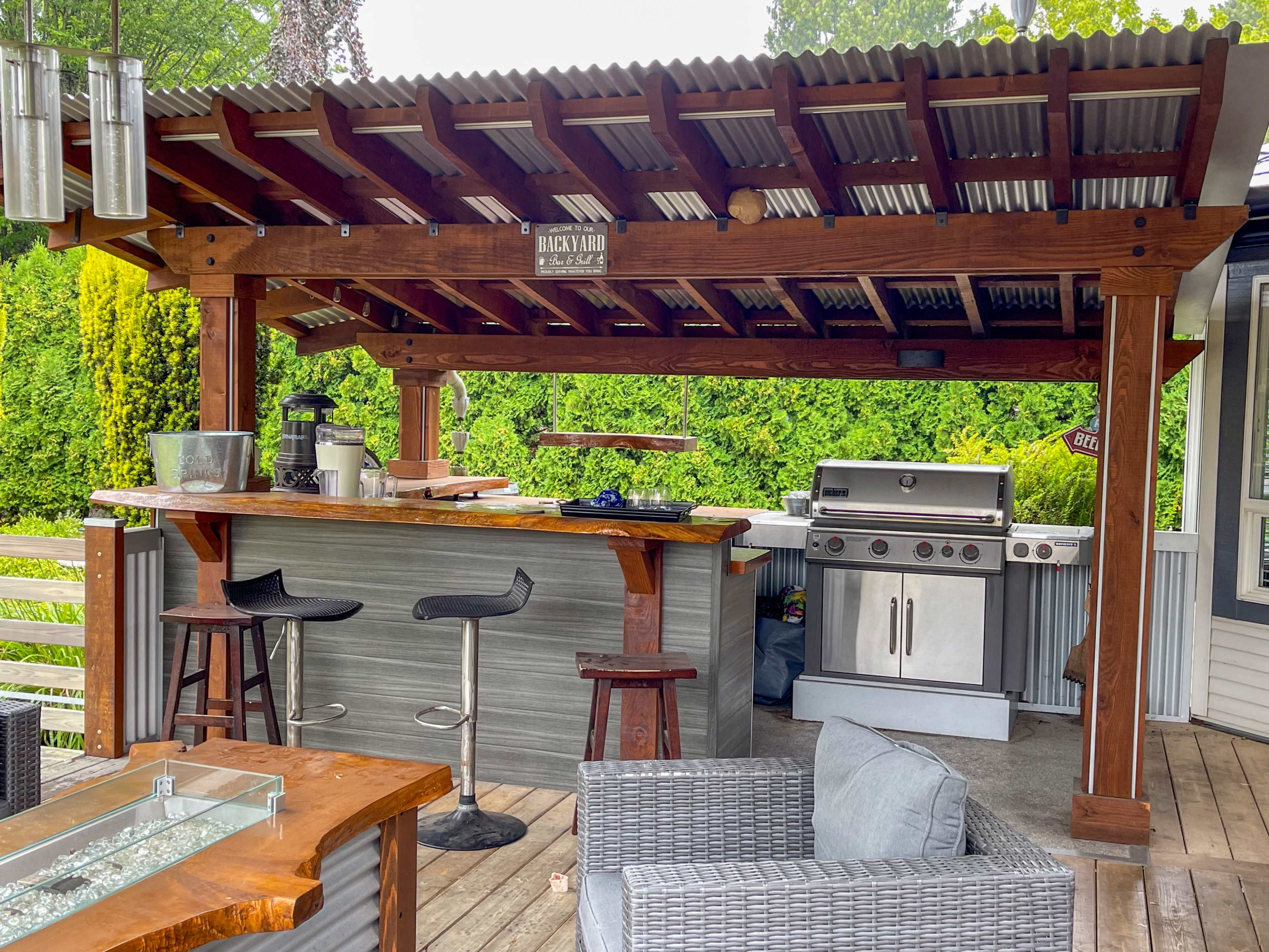 An outdoor kitchen area with a wooden pergola, featuring a bar with stools, a grill, and a countertop, surrounded by greenery.